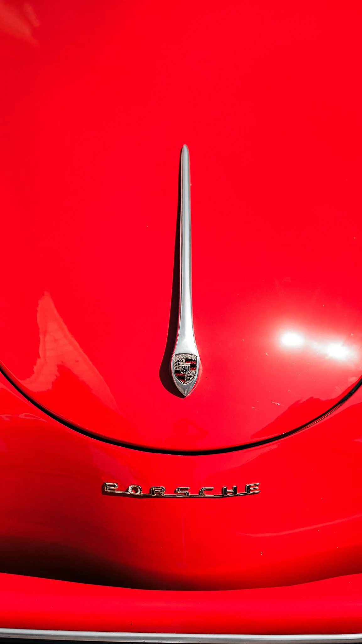 Close-up of a red Porsche vehicle hood with a chrome hood ornament featuring the Porsche emblem and the word 'Porsche' badge.