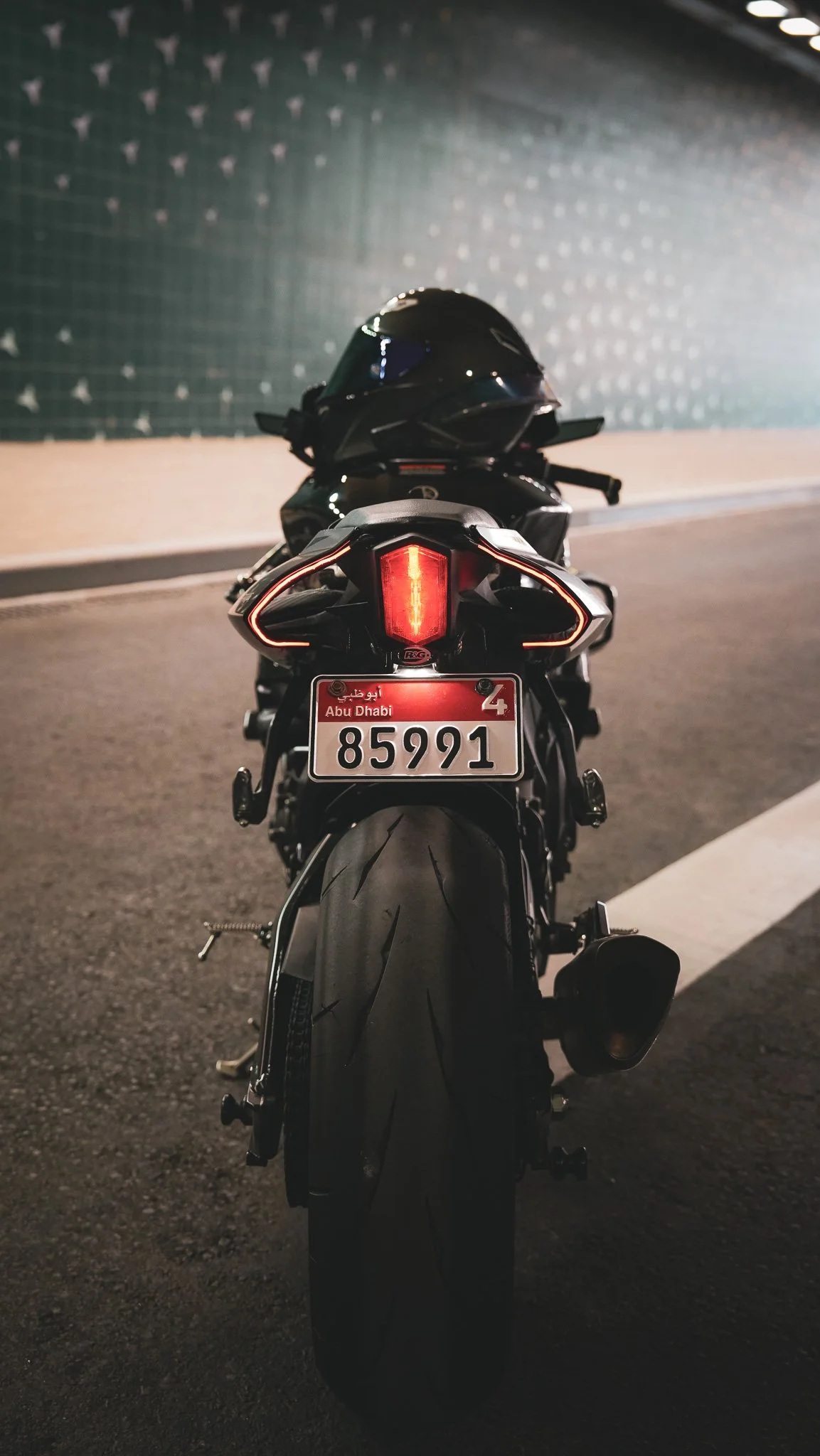 Rear view of a black motorcycle with a helmet resting on the seat, parked on a road at night. The motorcycle has a license plate from Abu Dhabi with the number 85991.