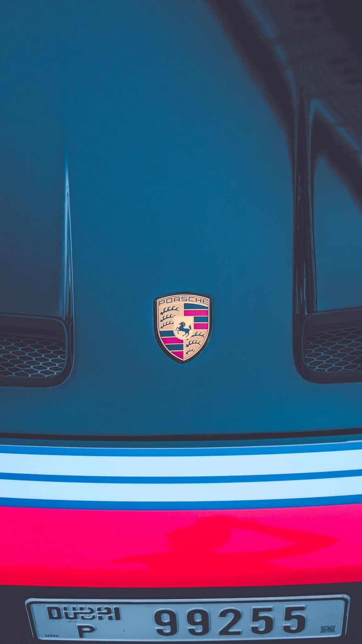 Front of a blue Porsche car with the Porsche emblem on the hood, a colorful stripe design below the emblem, and a license plate with the number 99255.