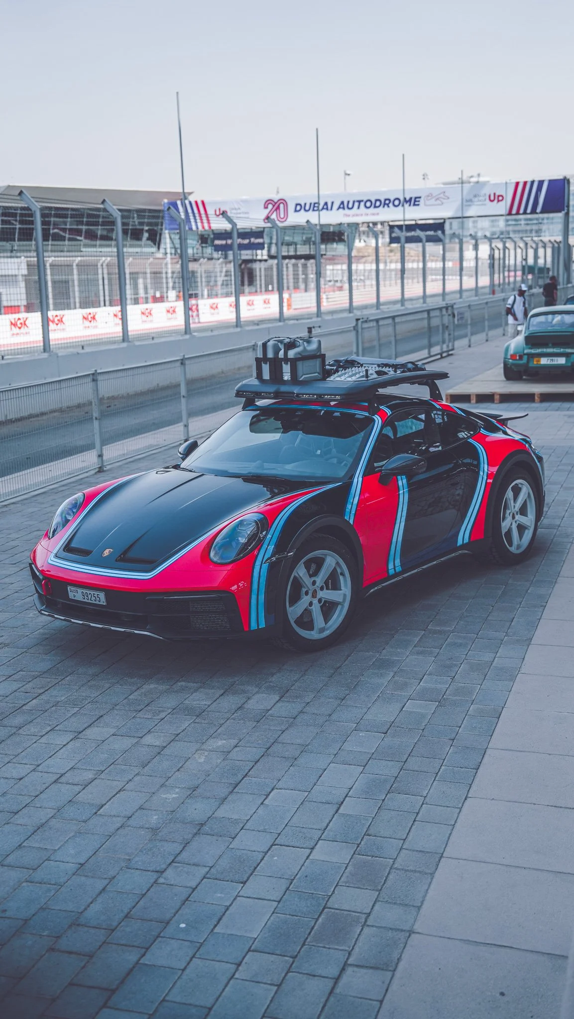 A race car with a red and black paint job and racing stripes is parked on a paved area near a race track at Dubai Autodrome. There is a background of fencing, race track, and a banner that reads 'Dubai Autodrome'.