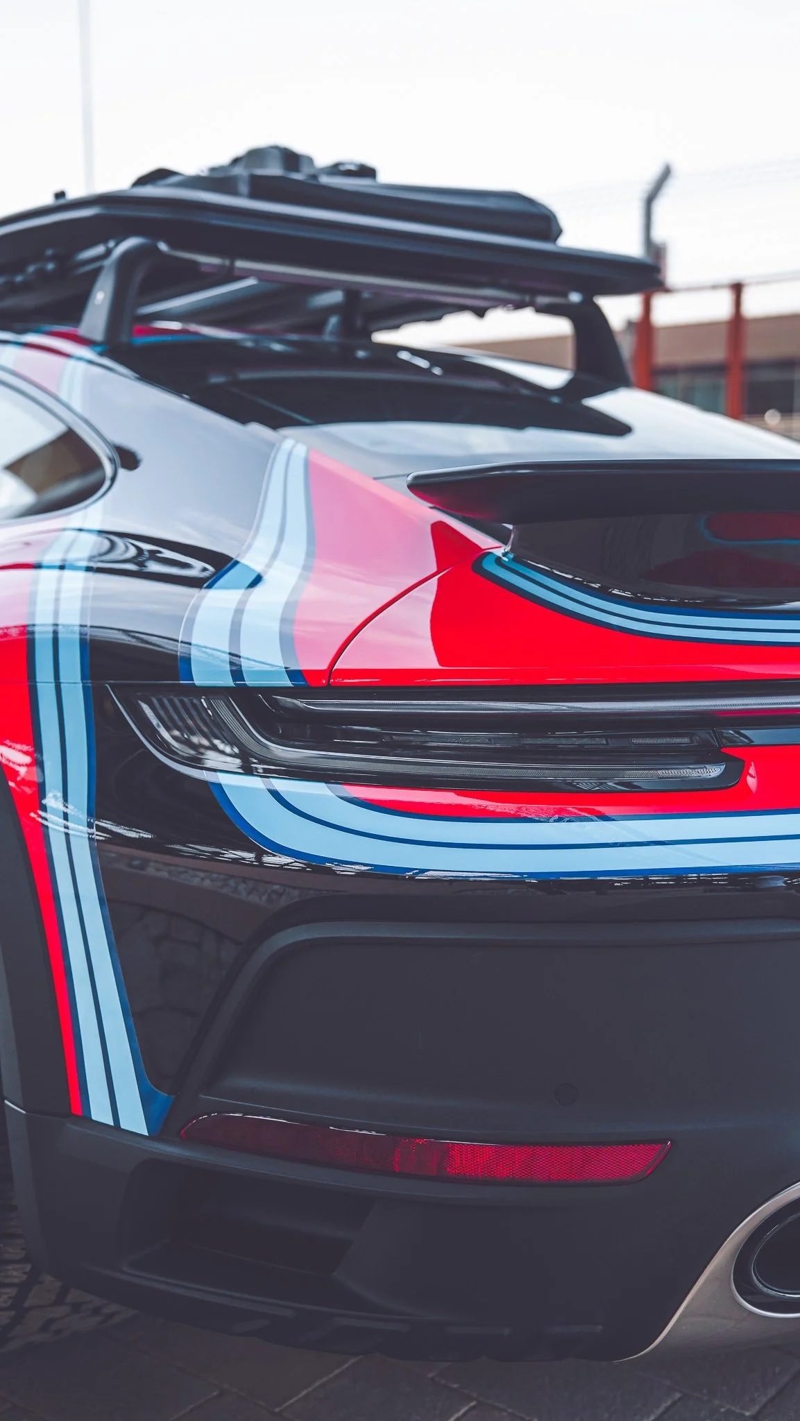 Close-up of the rear side of a racing car with a black, red, and blue striped paint job, featuring a prominent rear spoiler and sleek headlight design.