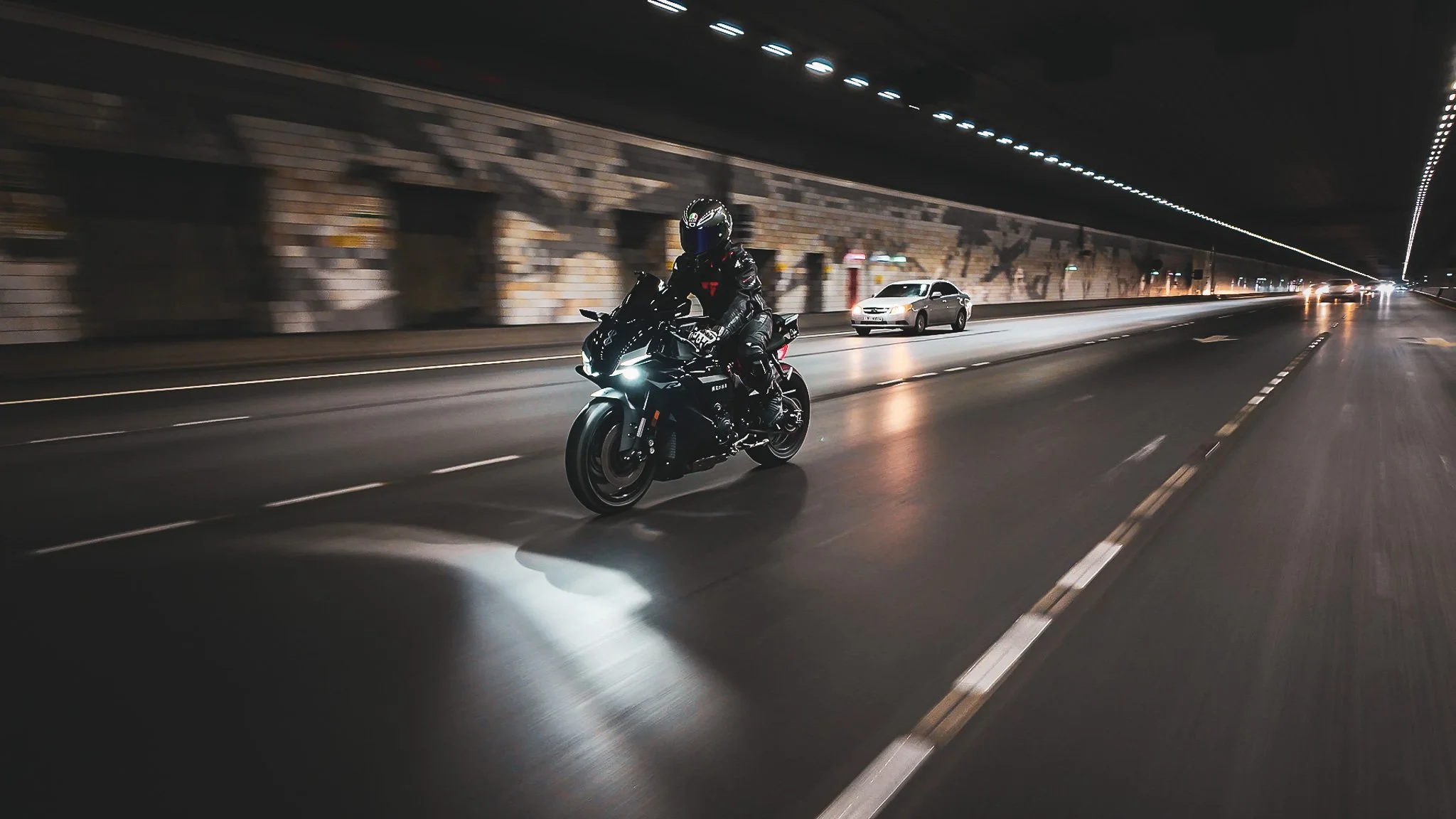 A motorcyclist in black gear and helmet riding a black motorcycle through a dimly lit tunnel, with a car behind on the road.