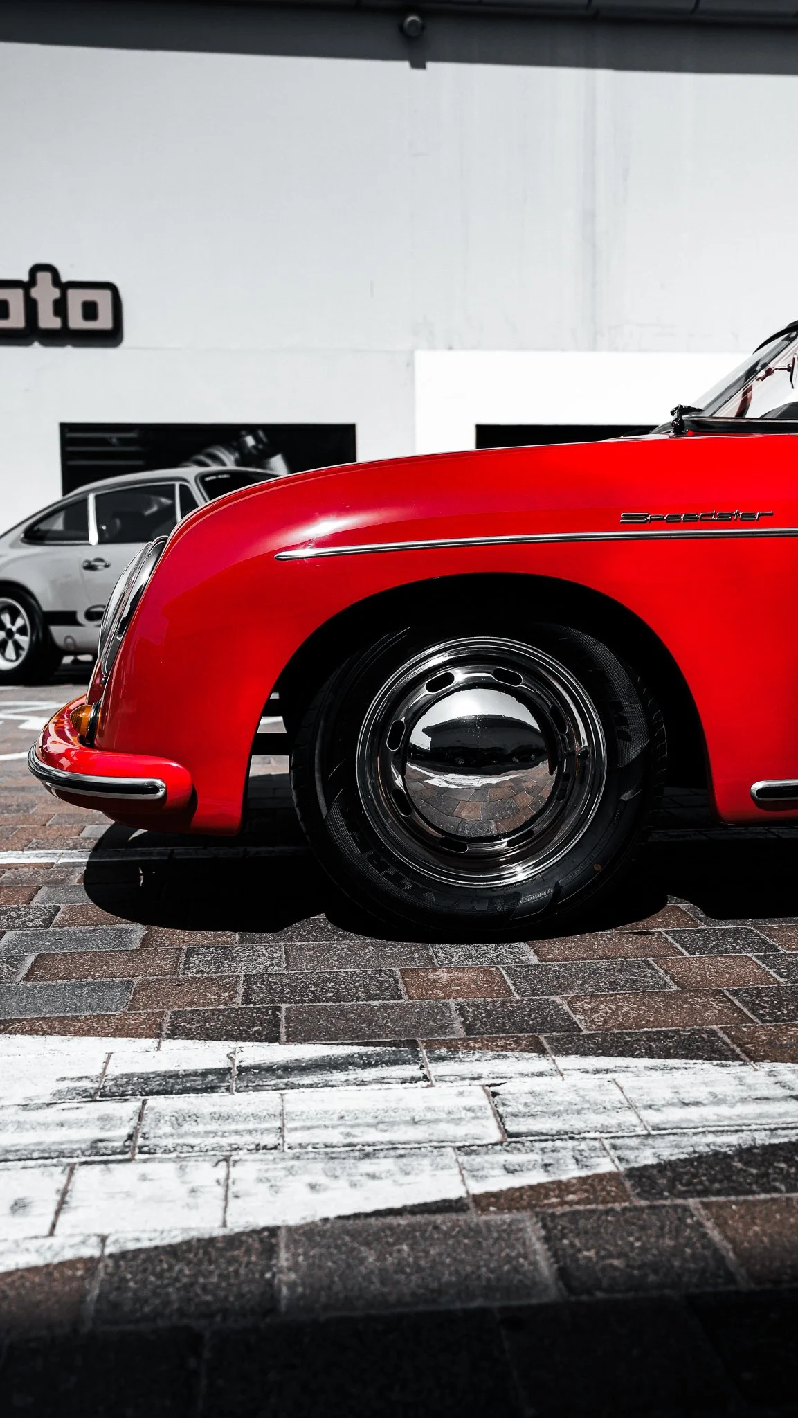 Close-up of a red vintage sports car with the word 'Speedster' on the side, parked on a brick surface. In the background, a gray car and part of a white building are visible.