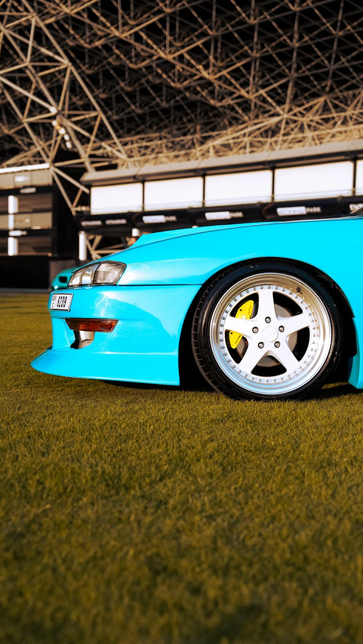 Close-up of a bright blue sports car with a white racing wheel and yellow brake caliper, parked on a grassy area at a racing stadium.