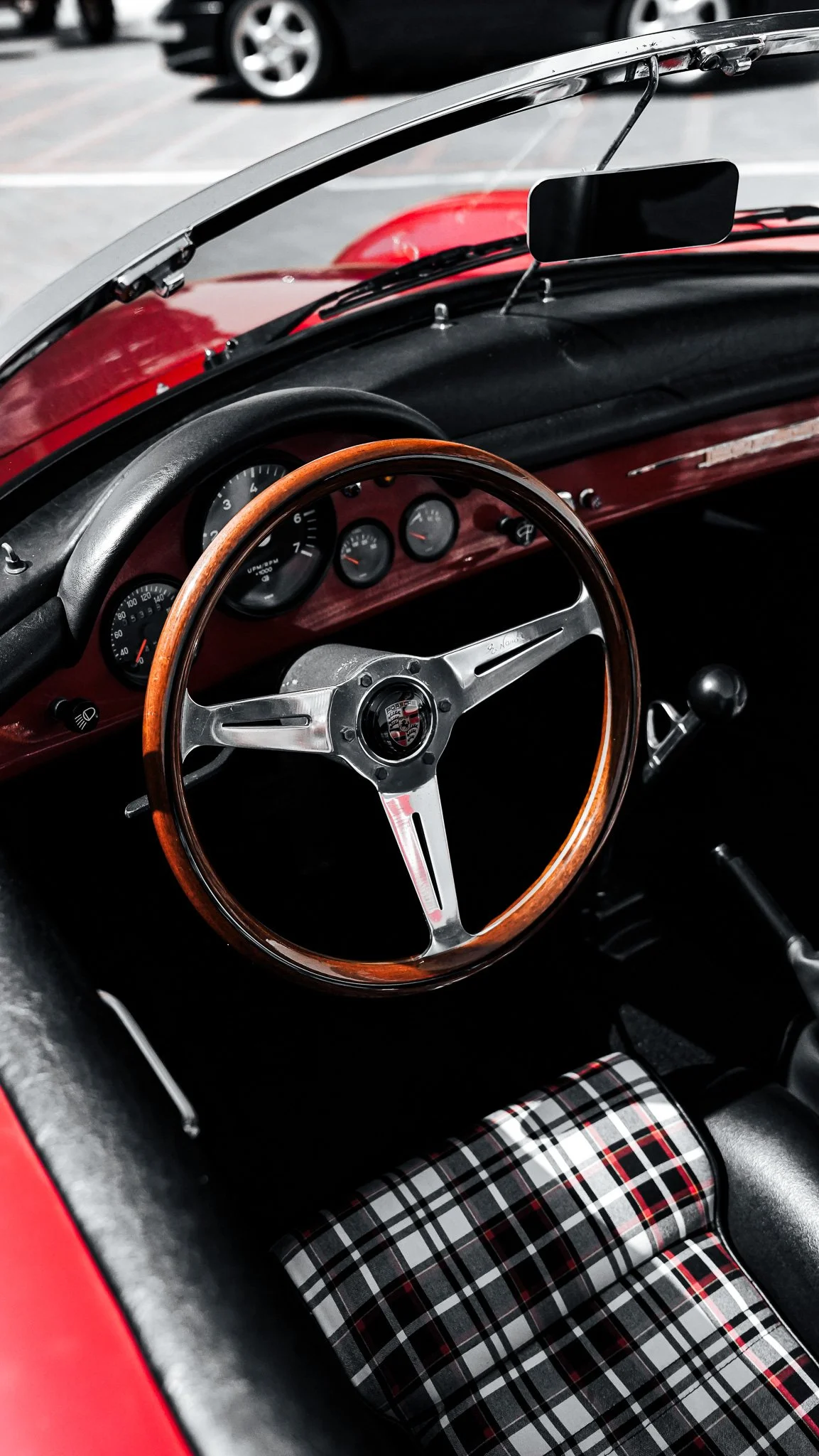 Interior of a classic red car showing a wood and metal steering wheel, several gauges on the dashboard, a rearview mirror, and plaid patterned seat upholstery.