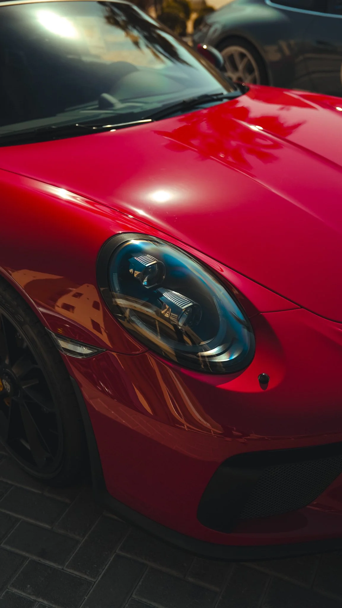 Close-up of a red sports car's front end, showing the headlight, hood, and part of the windshield with a reflection of the sky and surroundings.