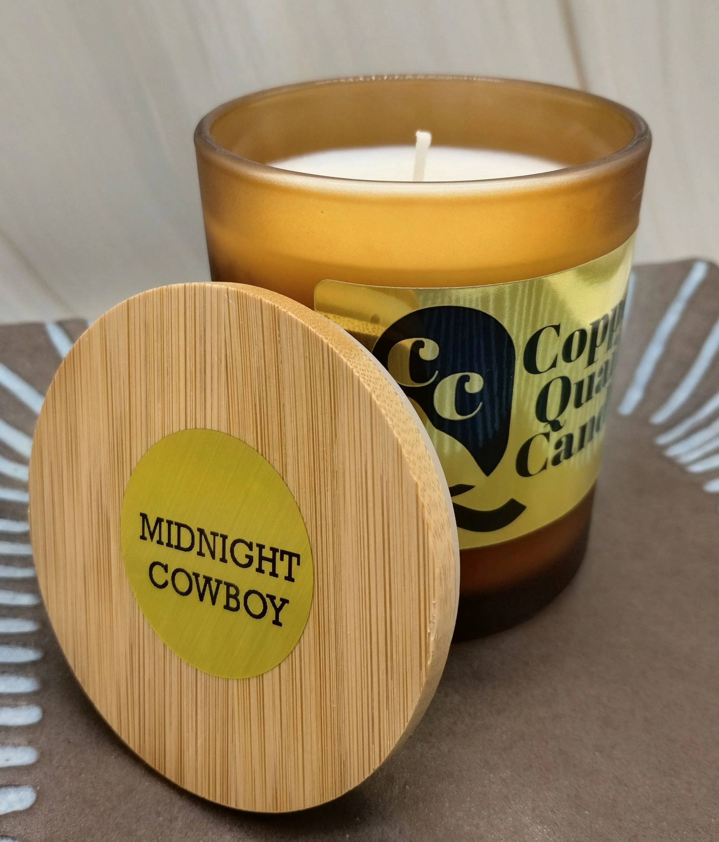Candle in an amber jar with a wooden lid leaned against it. Hand-poured soy candle. Midnight Cowboy fragrance.