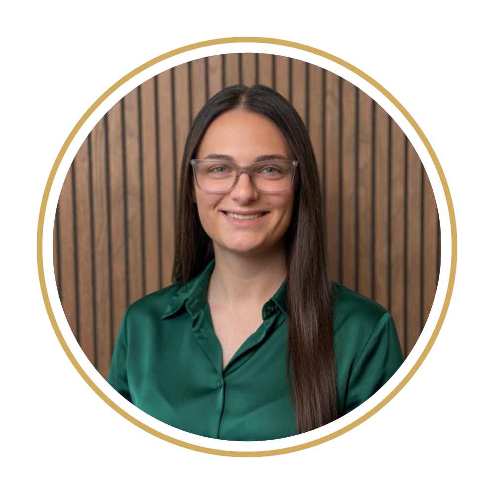 Ashley, wearing a dark green satin blouse and glasses, standing in front of a wooden panel background, smiling at the camera.