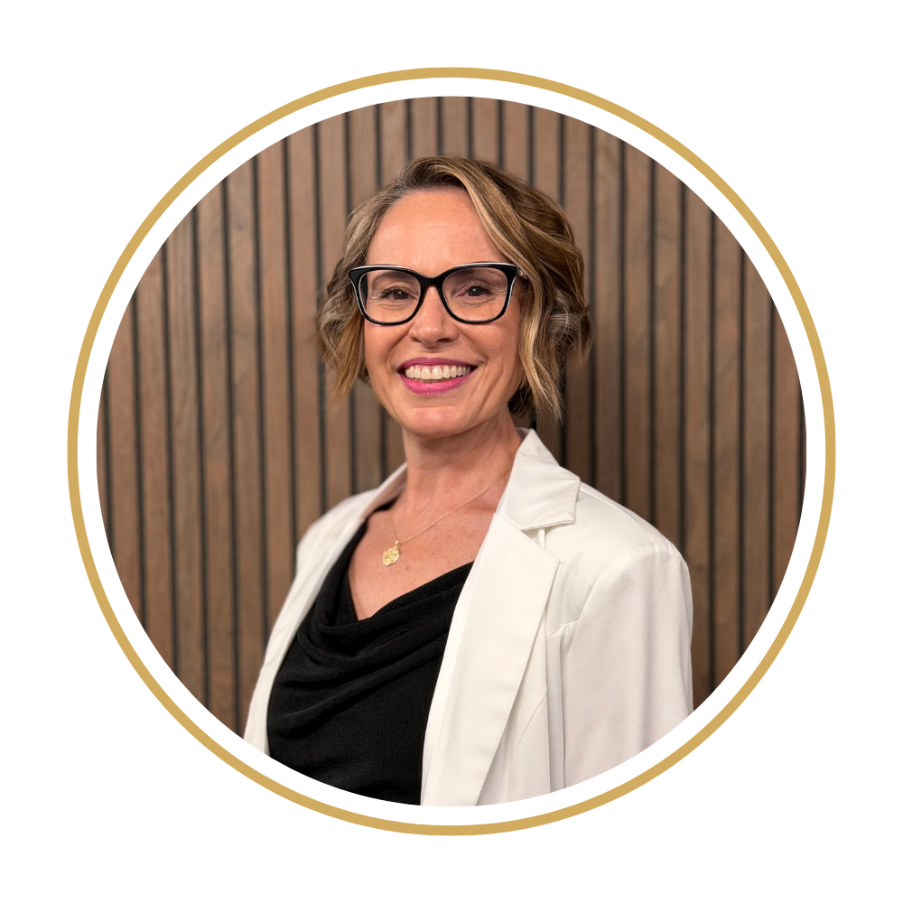 Leah wearing a white blazer with glasses, and a black top, standing in front of a wooden wall.