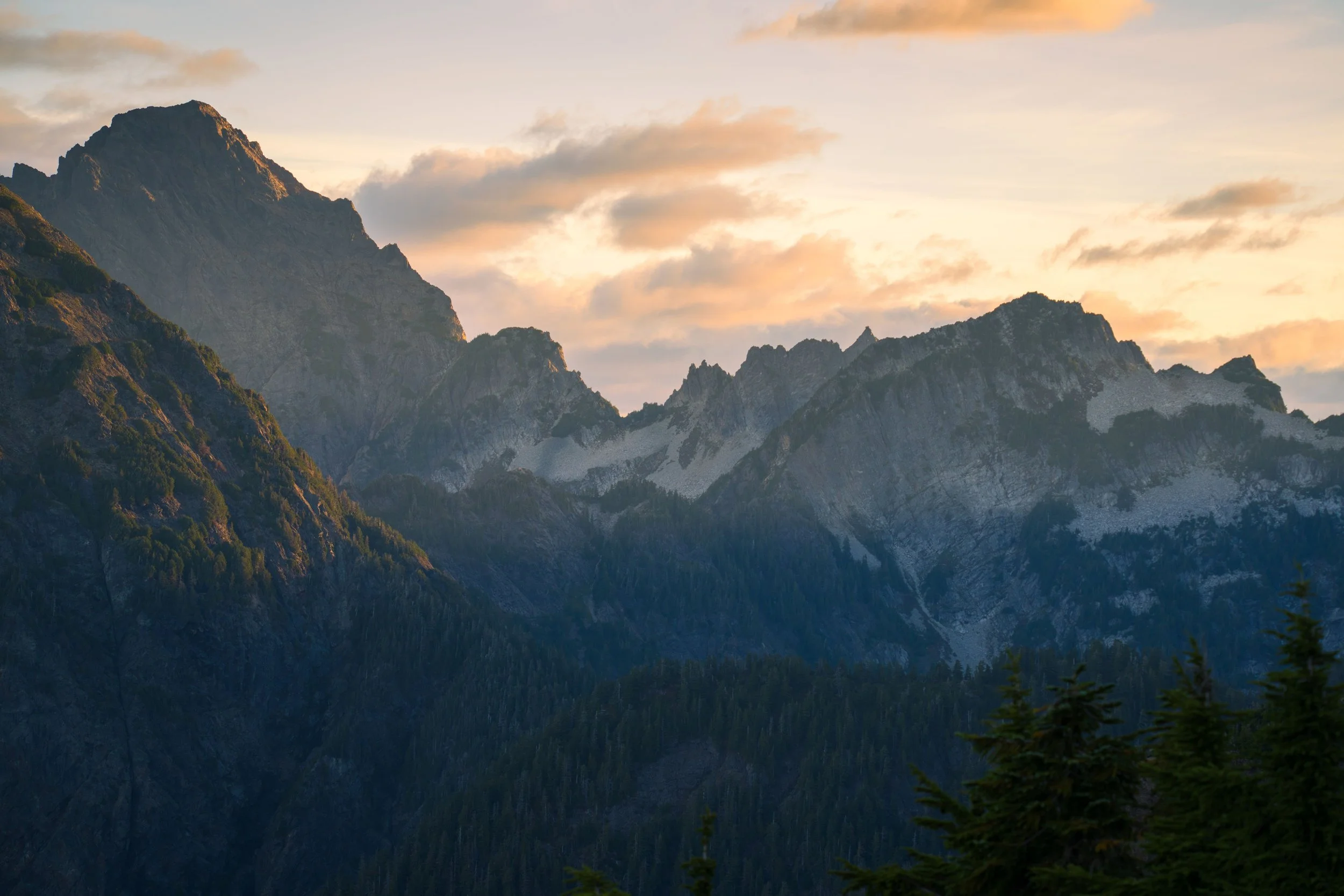 Mountain range with peaks, forested slopes, and a sky with clouds during sunset.