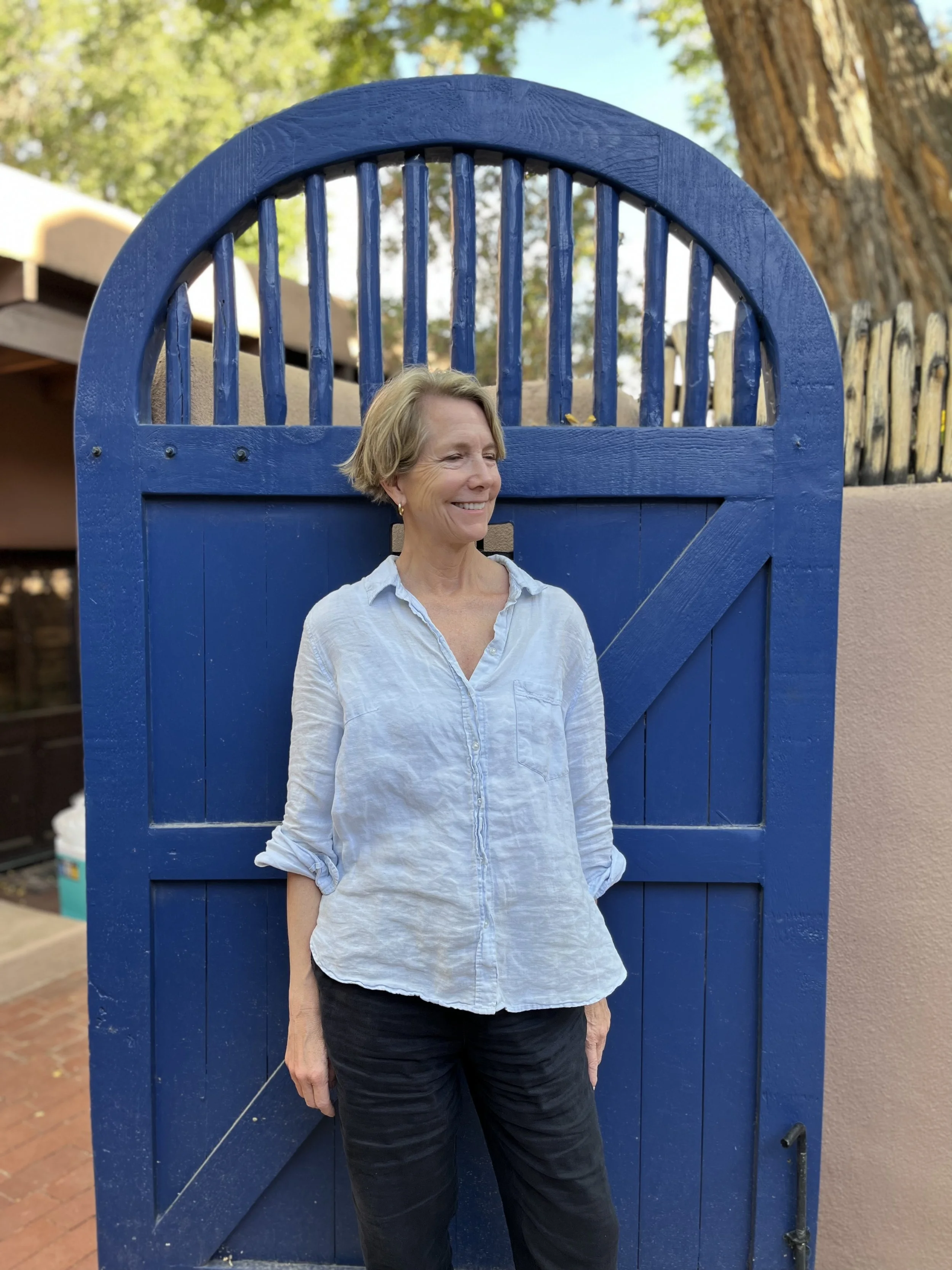 A woman with short blonde hair wearing a light blue button-up shirt and black pants standing in front of a decorative blue wooden gate with a curved top, outdoors on a sunny day with green trees in the background.