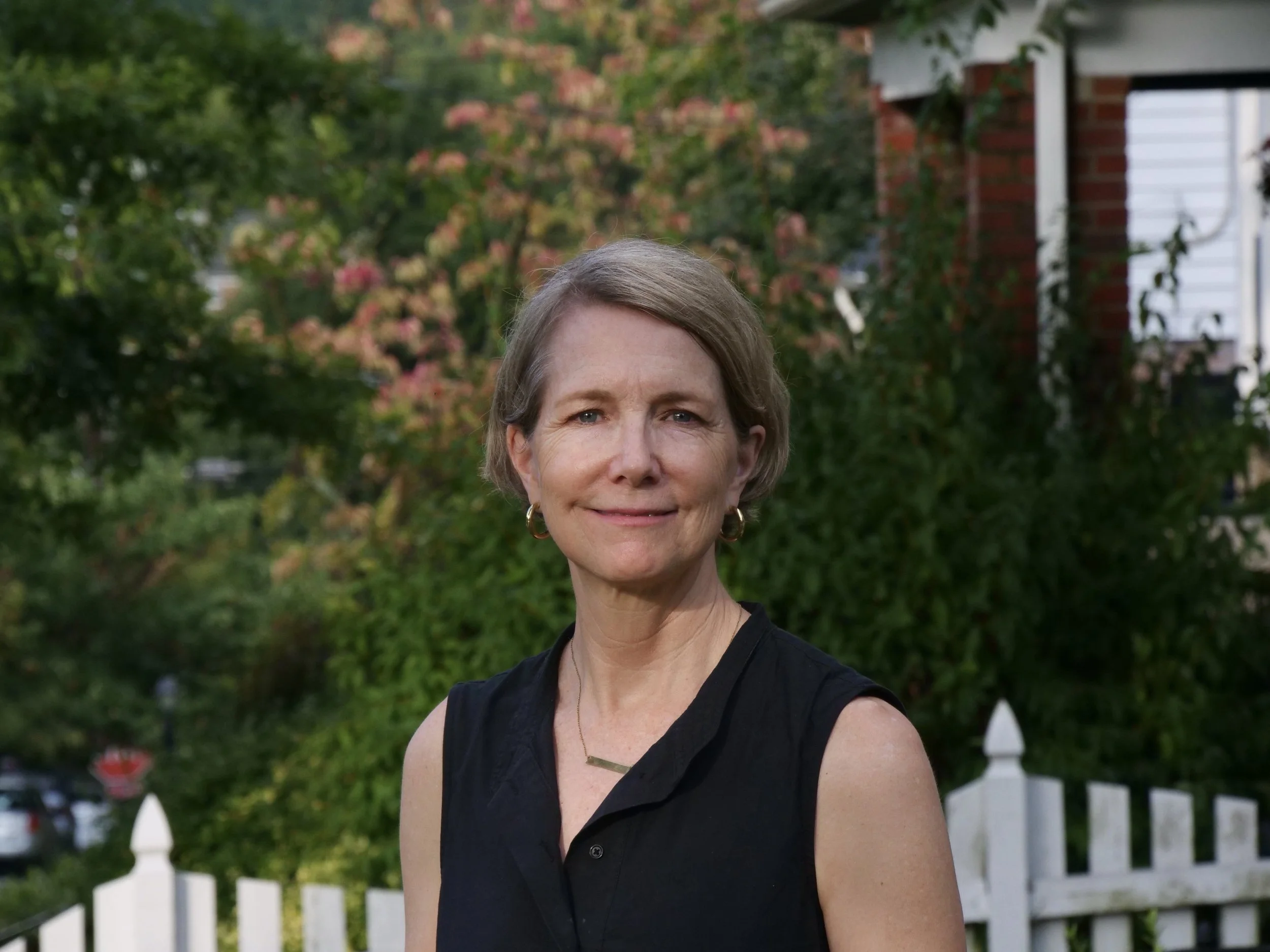 A woman with short, gray hair and earrings wearing a black sleeveless top, standing outdoors in front of a white picket fence, green trees, and a house with brick and white siding.