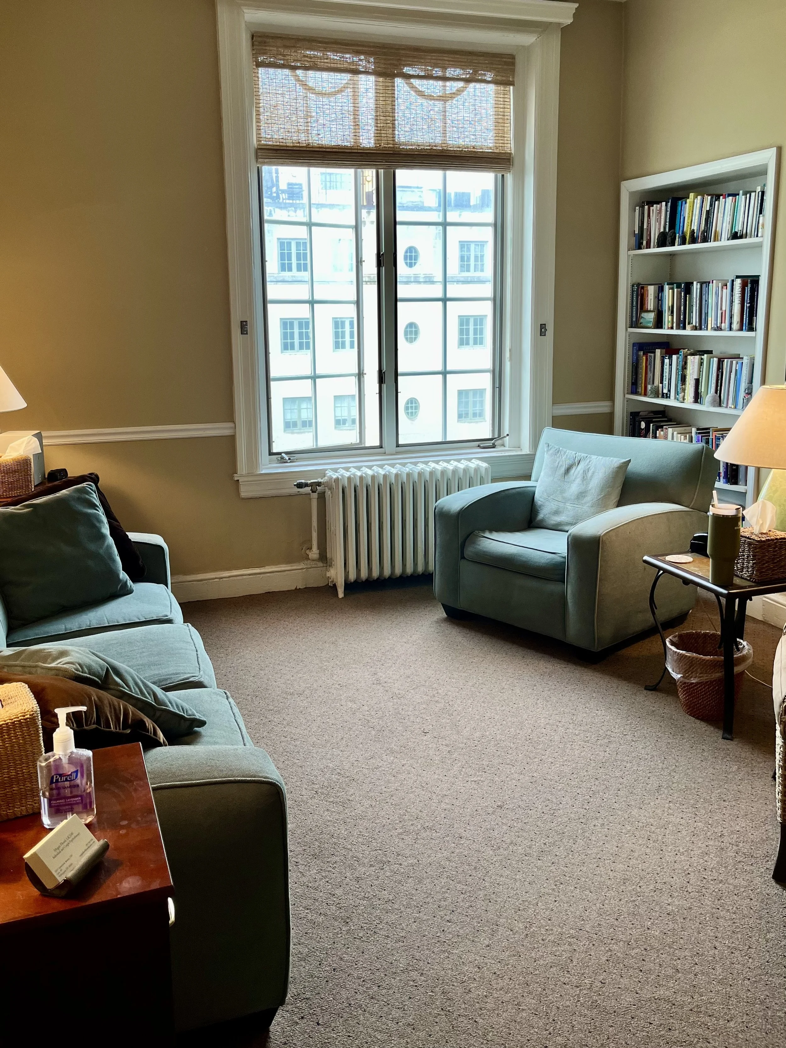 A cozy room with beige walls, featuring a large window with a bamboo shade, a blue armchair with a pillow, a bookshelf filled with books, a table with a lamp and decorations, and a couch with cushions. There is a radiator below the window and a side table with hand sanitizer and a sign.