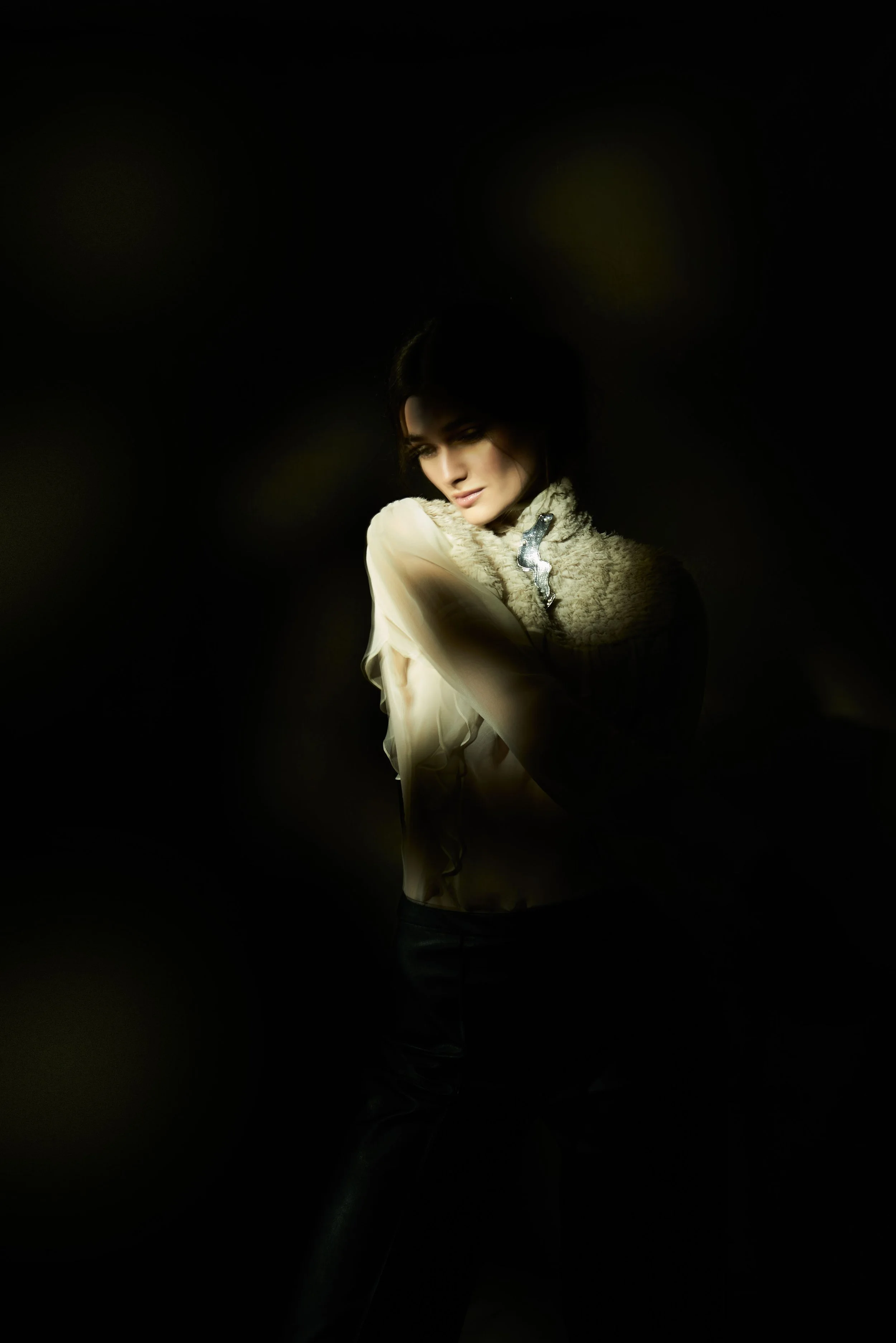 A woman with dark hair wearing a cream-colored jacket with a fur collar, standing in a dark setting with soft lighting highlighting her face and upper body, wearing a silver and diamond brooch on her shirt collar.