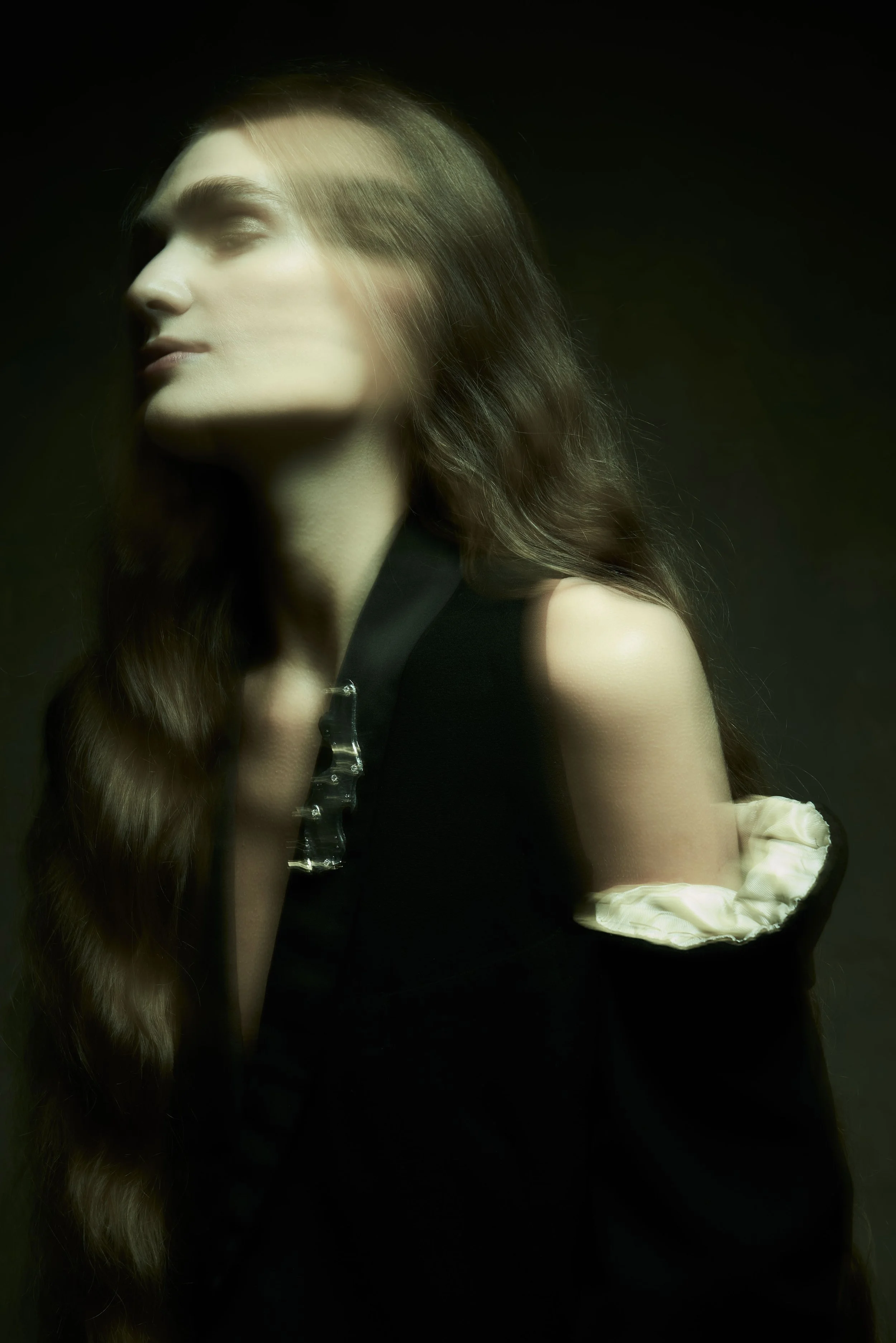 A woman with long, wavy brown hair and fair skin, wearing a black sleeveless top with ruffled cream-colored sleeves, wearing a silver brooch on the jacket lapel.