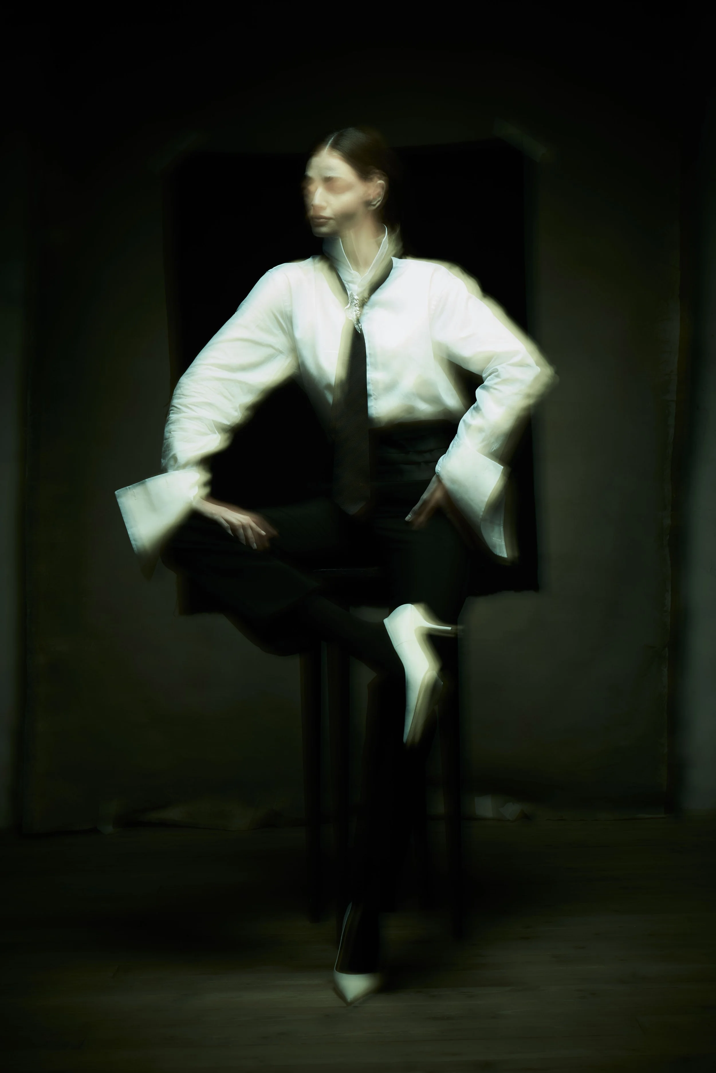 A woman in formal attire, with a glossy white shirt, black tie, and black pants, is seated on a chair in a dark room, with her hands on her hips and looking away from the camera.