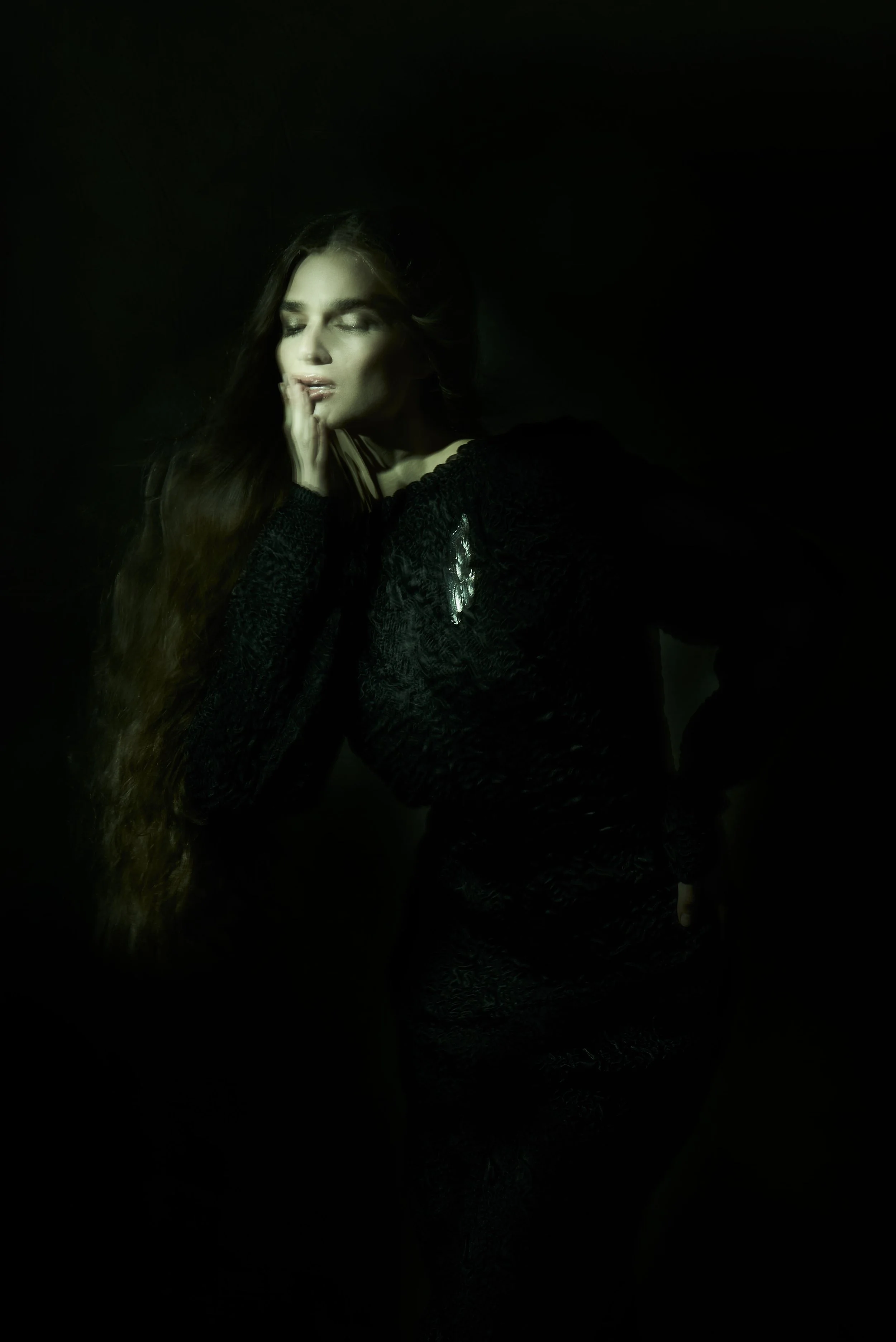 A woman with long, wavy brown hair closed eyes, touching her face with her right hand, wearing dark clothing, against a dark background.