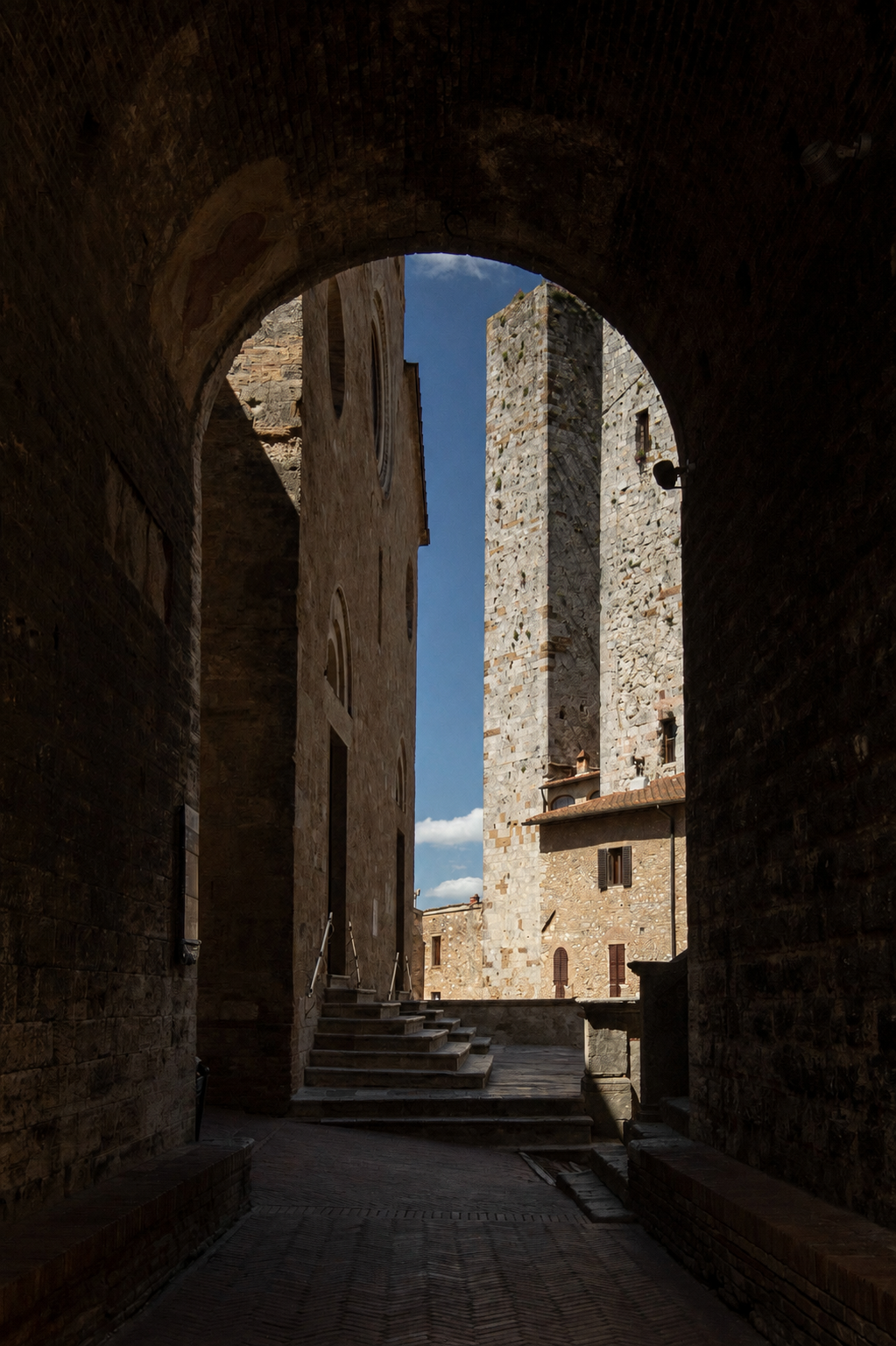 View of historic stone buildings and a tower taken through a dark stone archway, with steps leading up toward the structures and a bright blue sky with clouds in the background.