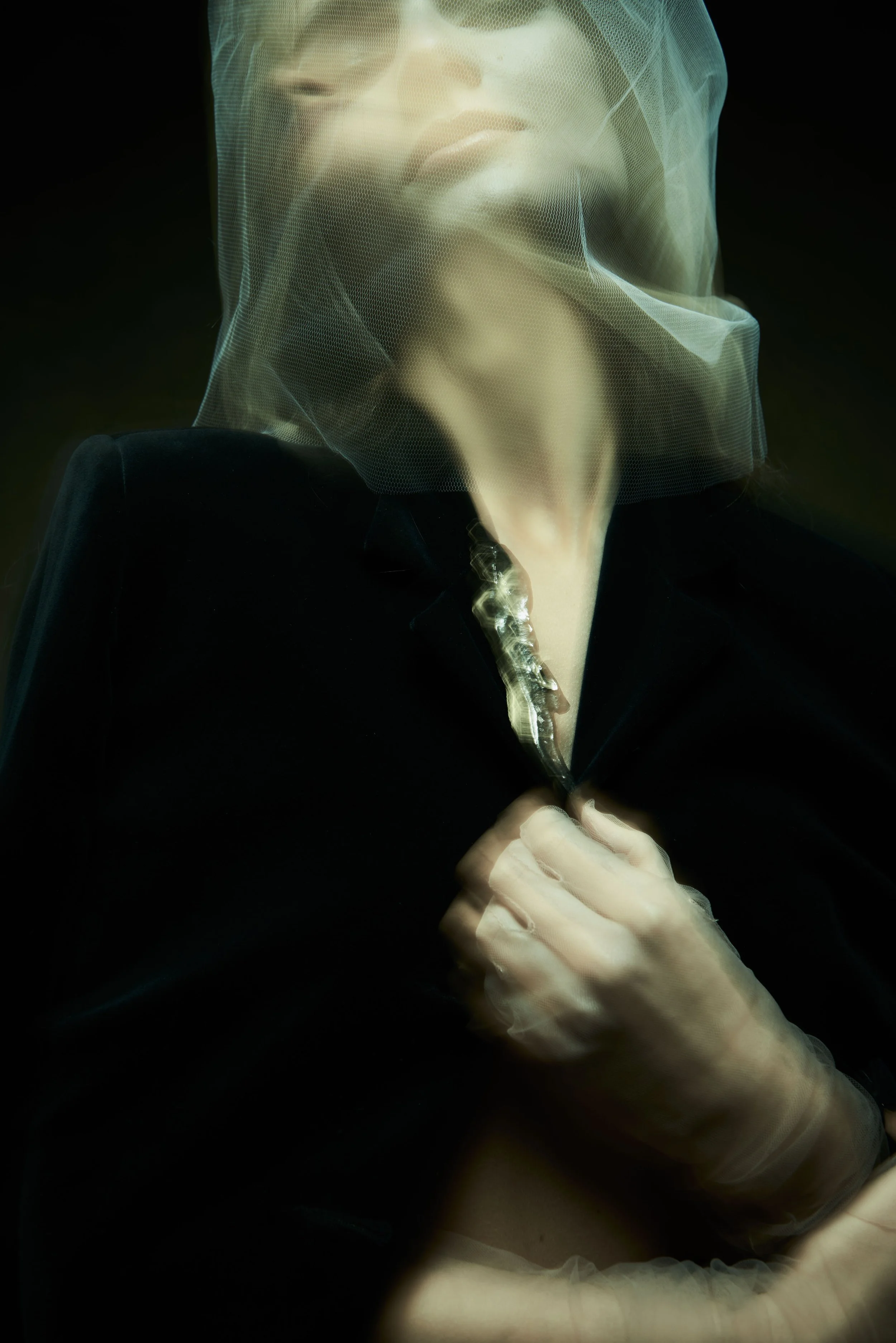 Person with a barrier of sheer fabric over their face, holding a long, curved object, with their hand visible at the bottom of the image, wearing a silver and diamond brooch on the jacket lapel.