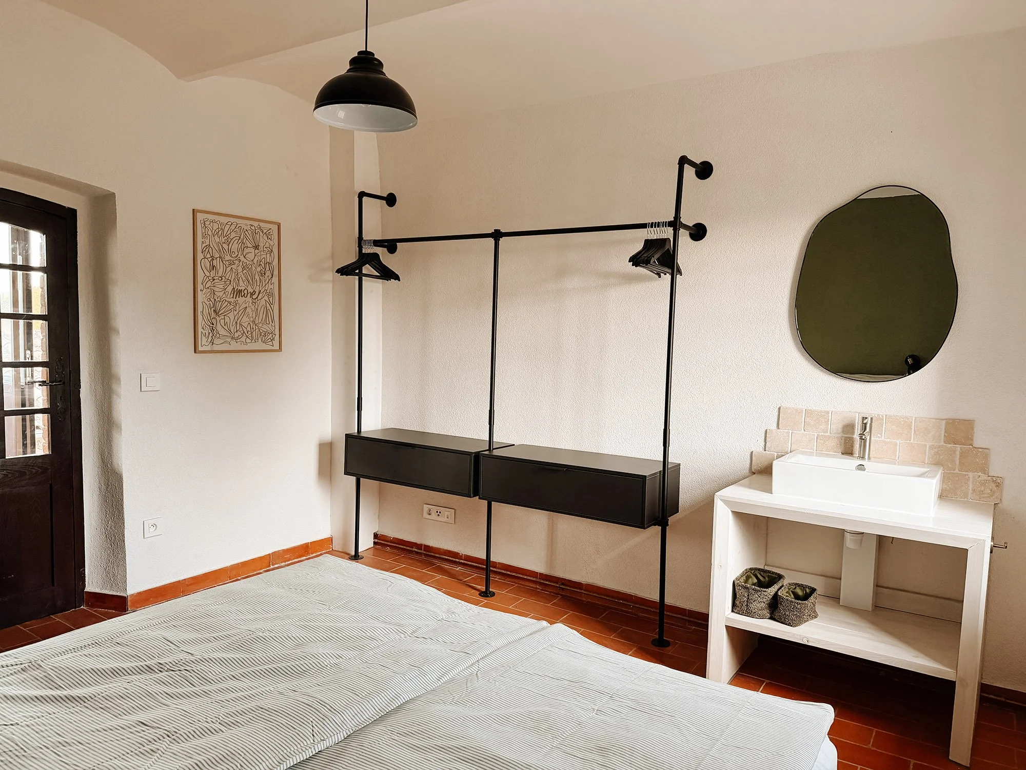 Ein Schlafzimmer mit weißem Bett, dunklem Kleiderschrank, Wanddeko, Spiegel und Waschbecken. Wand und Boden sind hell, die Decke ist leicht gewölbt.
