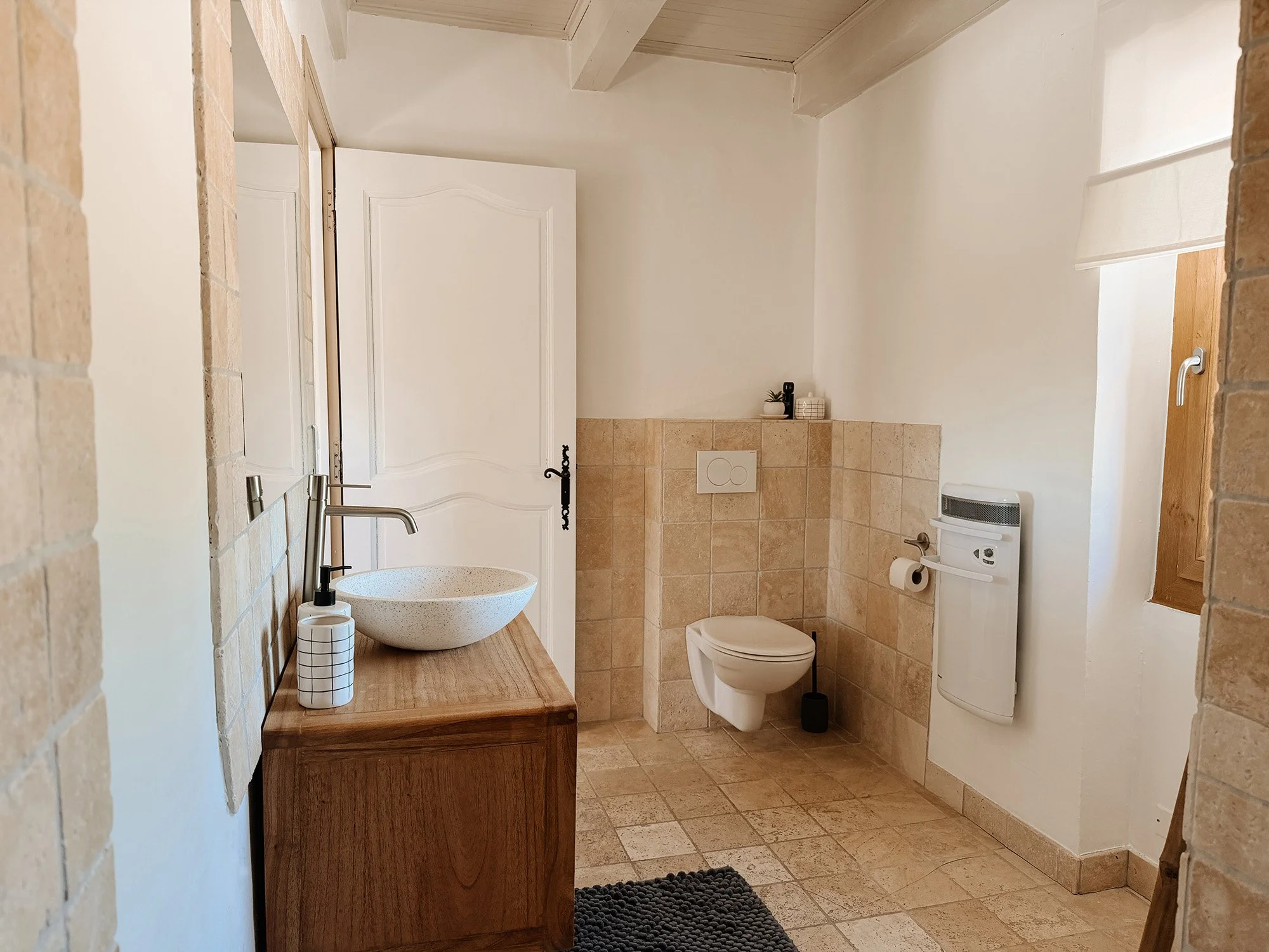 Ein Badezimmer mit weißer Wand, hellbraunen Fliesen, einem Wand-WC, einem Heizgerät, einem Waschtisch mit Waschbecken und einem Spiegel.