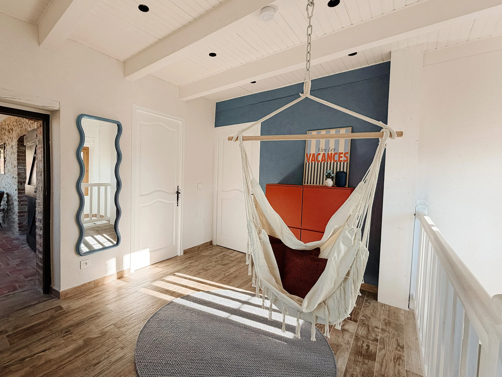 Ein heller Raum mit Holzfußboden, weißen Wänden und einer weißen Holzdecke. Im Raum hängt eine hängende Hängematte, und an der Wand befindet sich ein rustikaler Wandspiegel. Es gibt eine rote Kommode mit Dekorationsgegenständen und einem Poster mit d