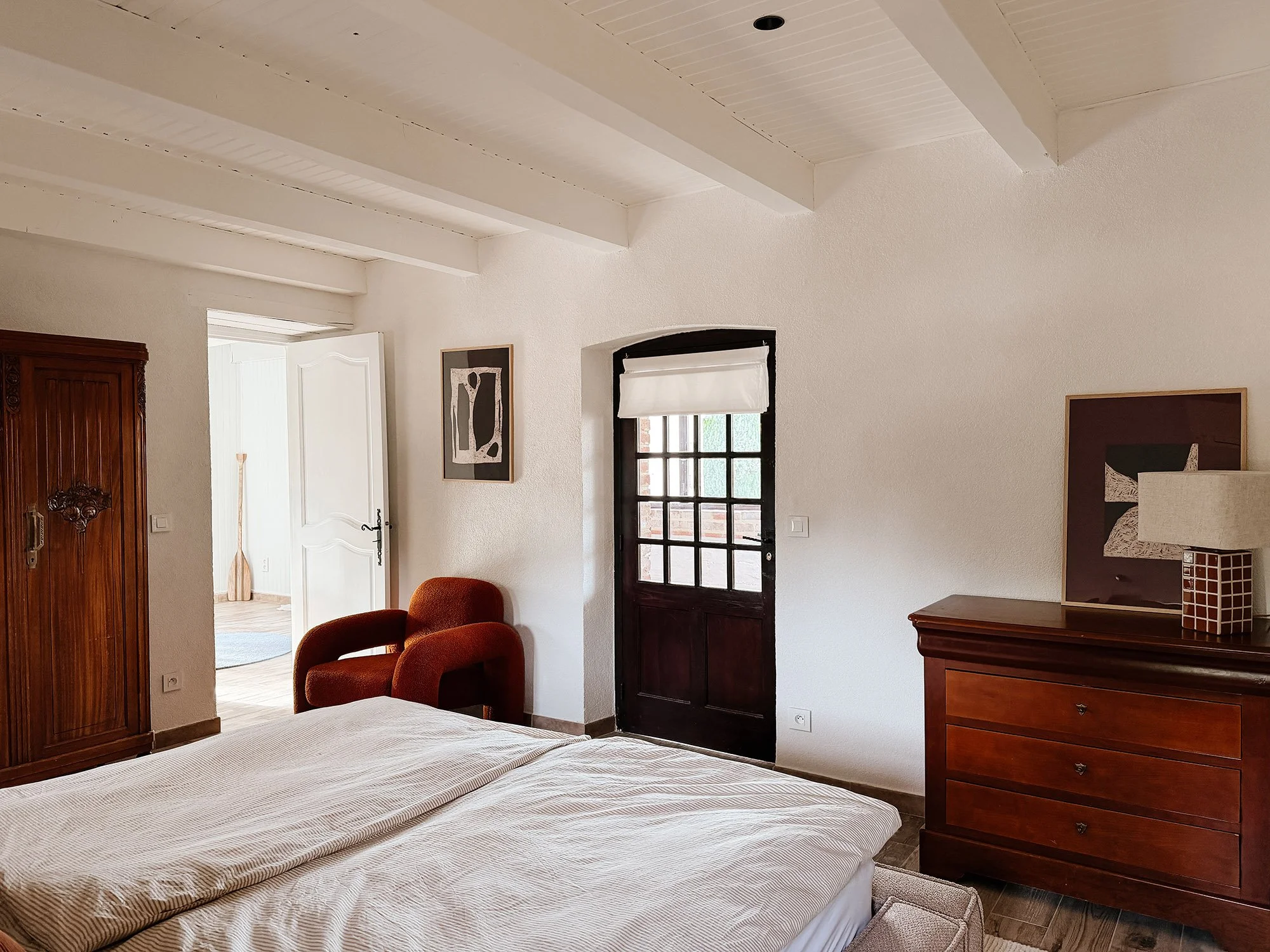 Ein Schlafzimmer mit einem Bett, einem Holzschrank, einem rot gepolsterten Stuhl, einem Wandbild, sowie einem Fenster mit weißen Vorhängen, und einer offenen Tür im Hintergrund.