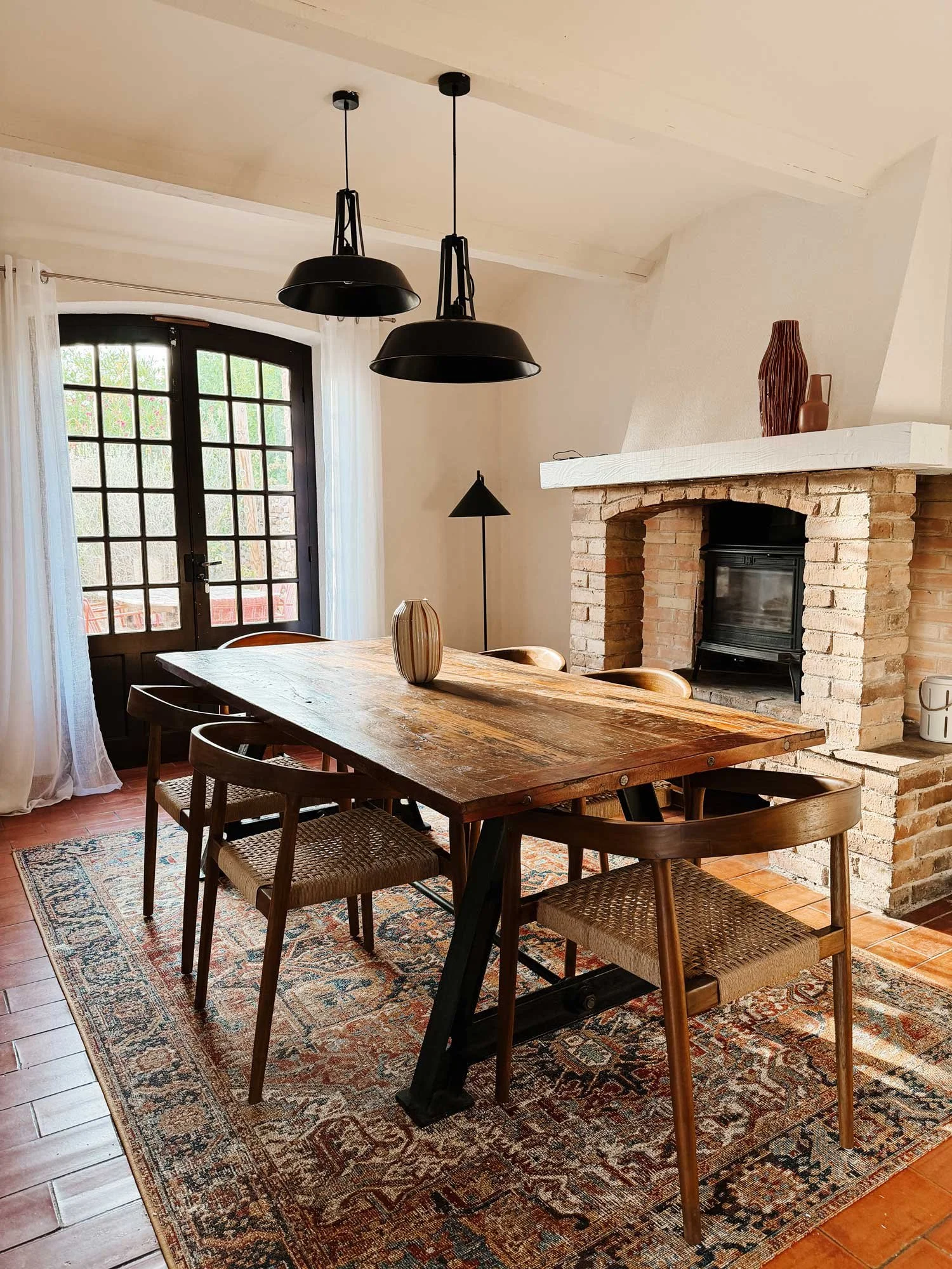 Der Raum ist ein Esszimmer mit einem großen Holztisch und mehreren Stühlen. Es gibt eine Wand mit einem Kamin, auf dem zwei Vasen stehen. Es gibt große Fenster mit weißen Vorhängen und einen Läufer mit Mustern auf dem Holzfußboden. Es hängen zwei schwarze Lampen von der Decke.