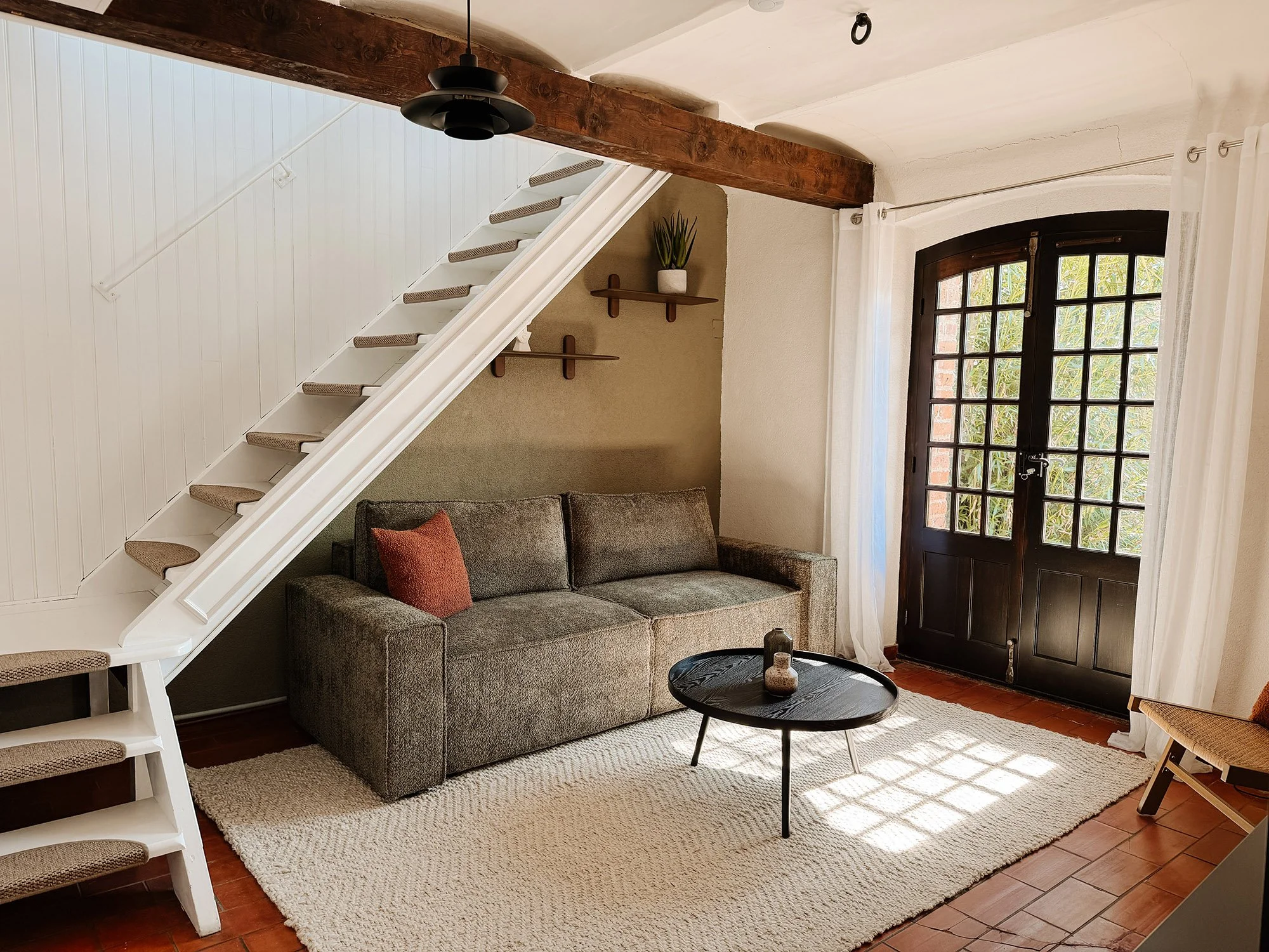 Ein Wohnzimmer mit einem grauen Sofa, einem schwarzen Kaffeetisch, einem weißen Teppich, einem großen Fenster mit weißen Vorhängen, einer Holzdecke, einem Holzträger, einer weißen Wand mit Wandregalen und Pflanze, an der rechten Seite eine Tür.