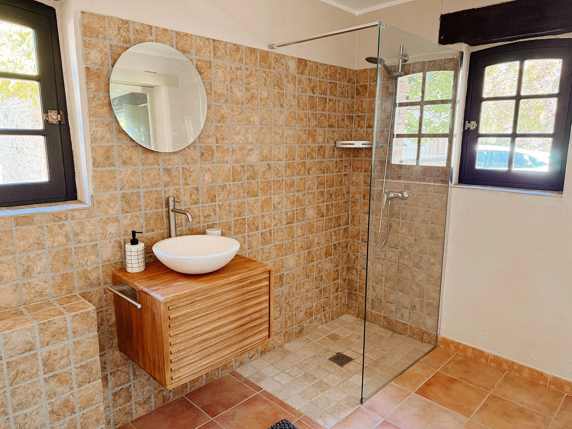 Ein Badezimmer mit gemusterten Fliesen, einem runden Waschbecken auf einem Holzunterschrank, einem Spiegel, einem Fenster und einer abgetrennten Dusche mit Glaswand.
