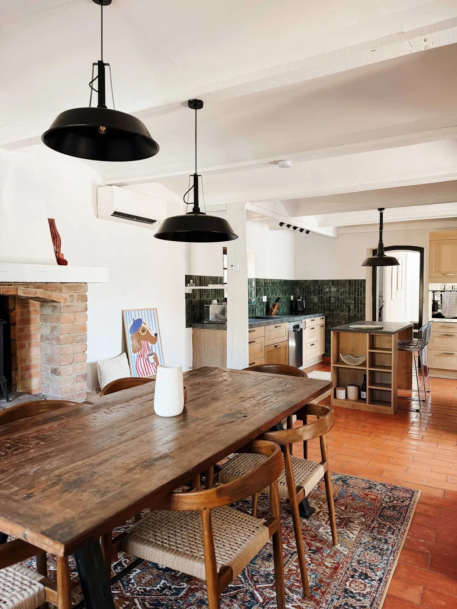 Ein heller Wohnraum mit einem großen Holztisch, mehreren Stühlen und schwarzen Deckenleuchten an der Decke. Im Hintergrund ist eine Küche mit grünen Fliesen und holzfarbenen Schränken sichtbar, sowie eine Wand mit einem Kamin und einem Kunstwerk mit 