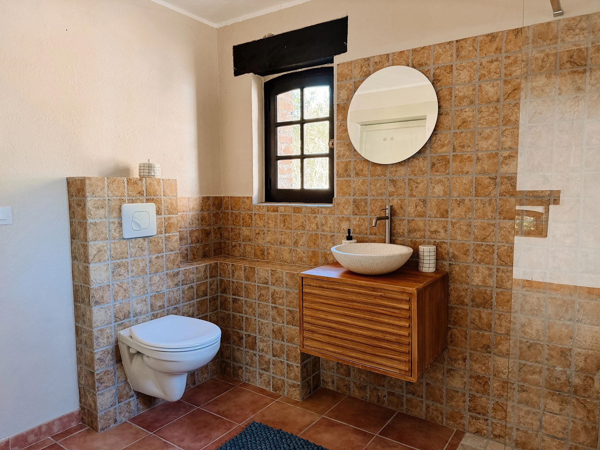 Ein Badezimmer mit einer weißen Toilette, einem runden Waschtisch, einer Holzkommode, einem runden Spiegel, einer Fensteröffnung, braun-orangen Fliesen und einem blauen Teppich.