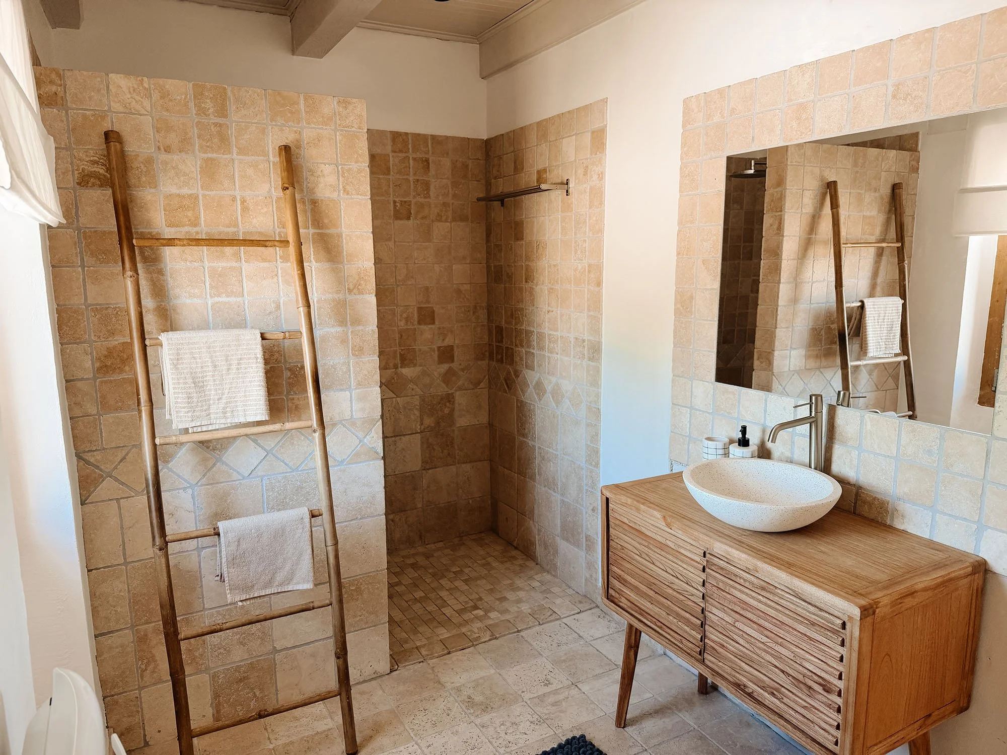Ein Badezimmer mit einer Dusche, einem Waschbecken und einer Holzkonsole. Es gibt einen Bambus-Trockengestell mit Handtüchern, Fliesen in Erdtönen und einen großen Spiegel.