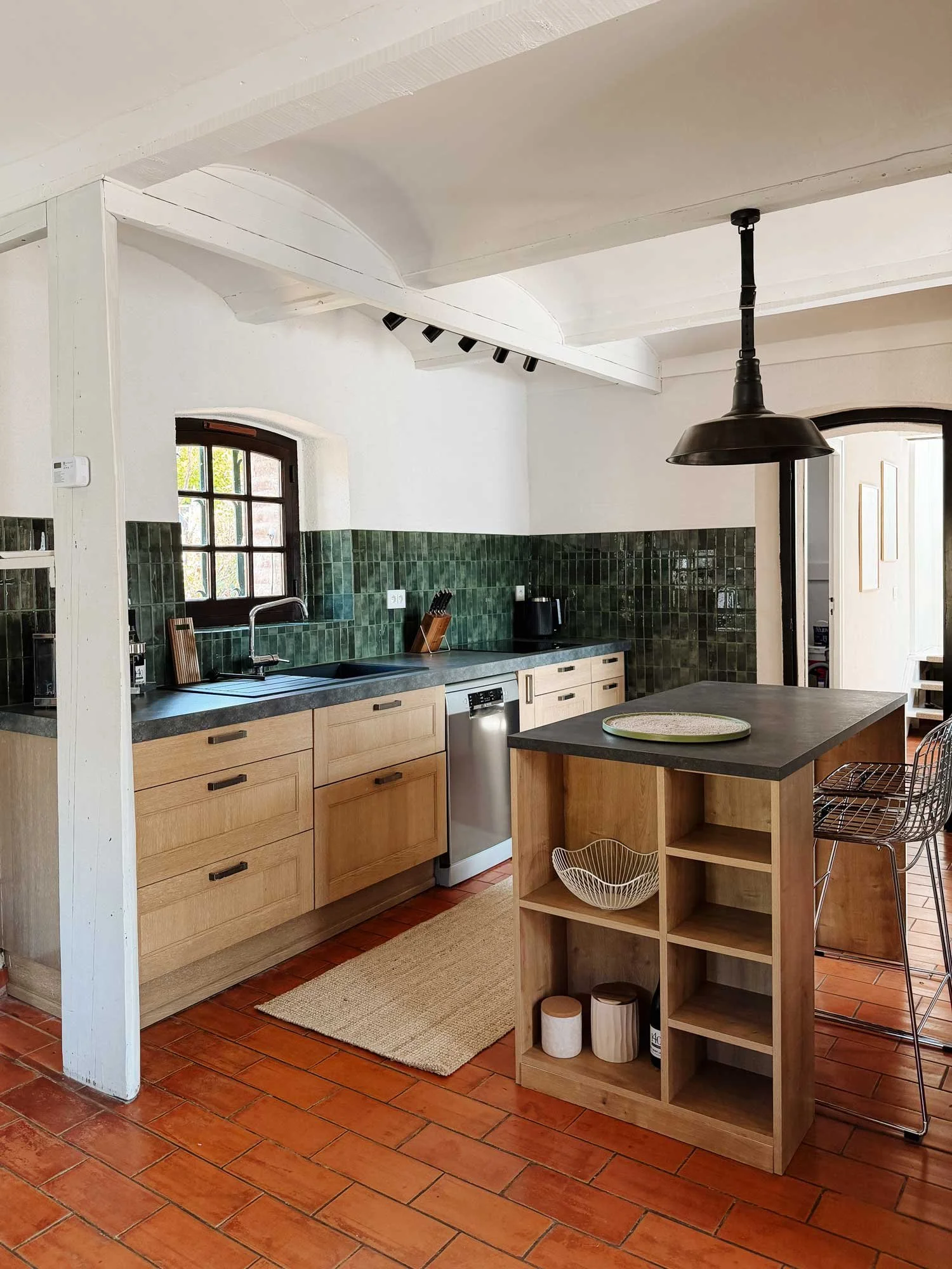 Moderne Küche mit grüner Fliesenrückwand, Holzschränken, Küchenspüle unter Fenster, zentraler Kücheninsel aus Holz, schwarze Hängeleuchte, offene Regale mit Küchenutensilien, terrakottafarbener Boden, natürliche Beleuchtung.