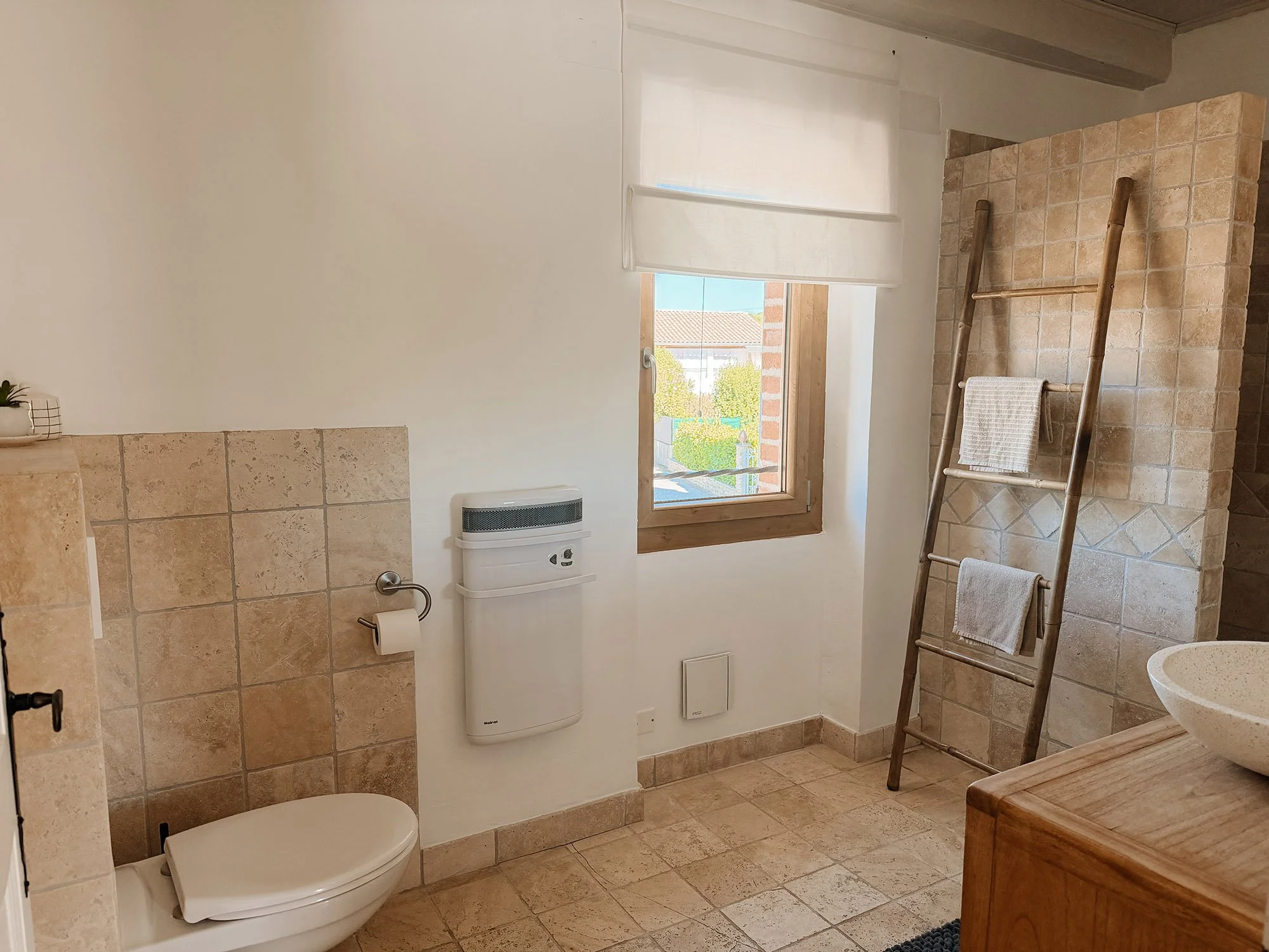 Ein Badezimmer mit einer Toilette, einem Heizkörper, einem Fenster mit Blick auf Häuser und Bäume, einem Holzregal mit Handtüchern und einer Wand aus beige Kacheln.