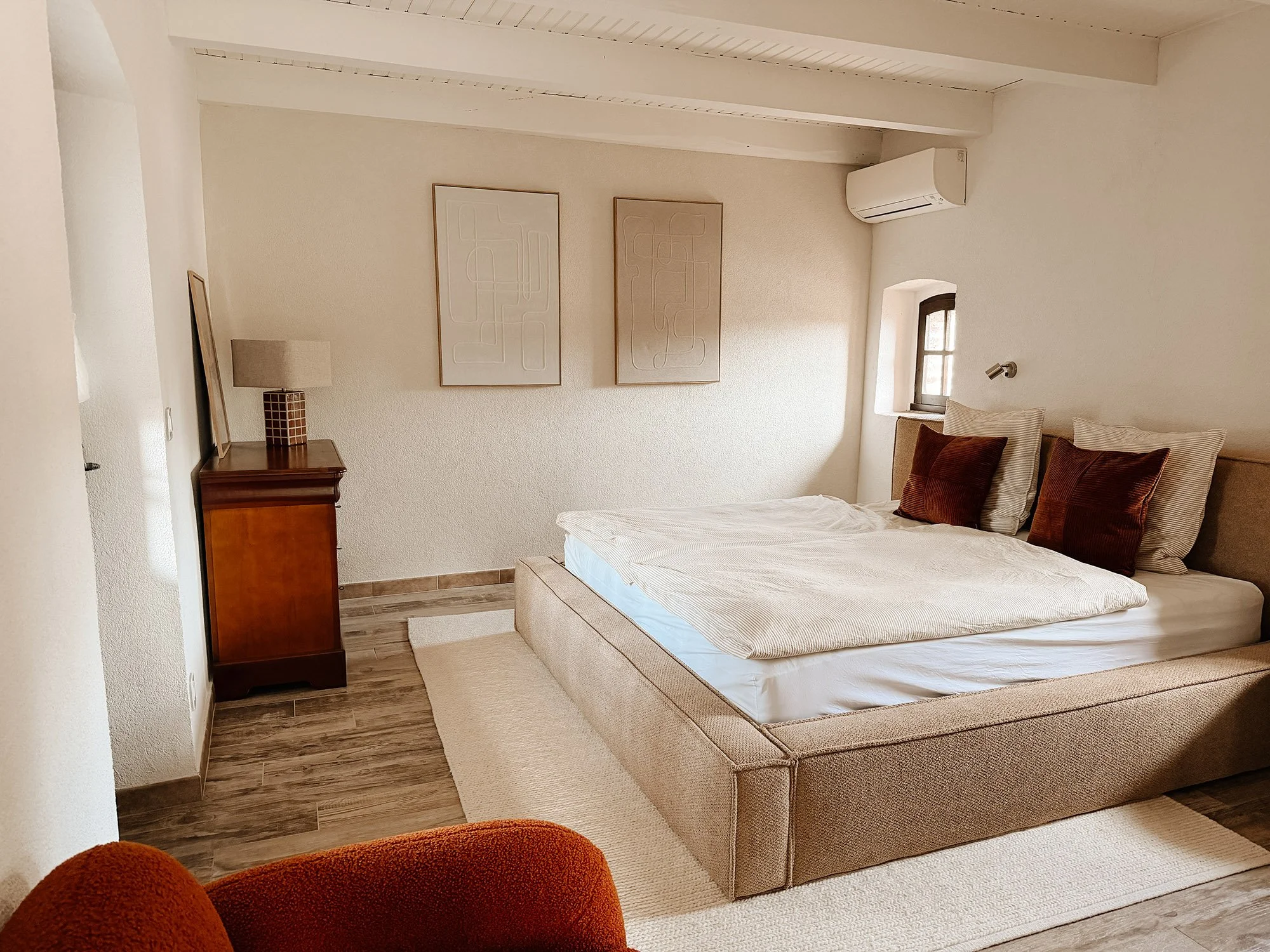 Das Bild zeigt ein Schlafzimmer mit einem Doppelbett, mehreren Kissen, einem kleinen Fenster, einer Nachttischlampe, Wanddekorationen, einem Kleiderschrank und einem Klimagerät.