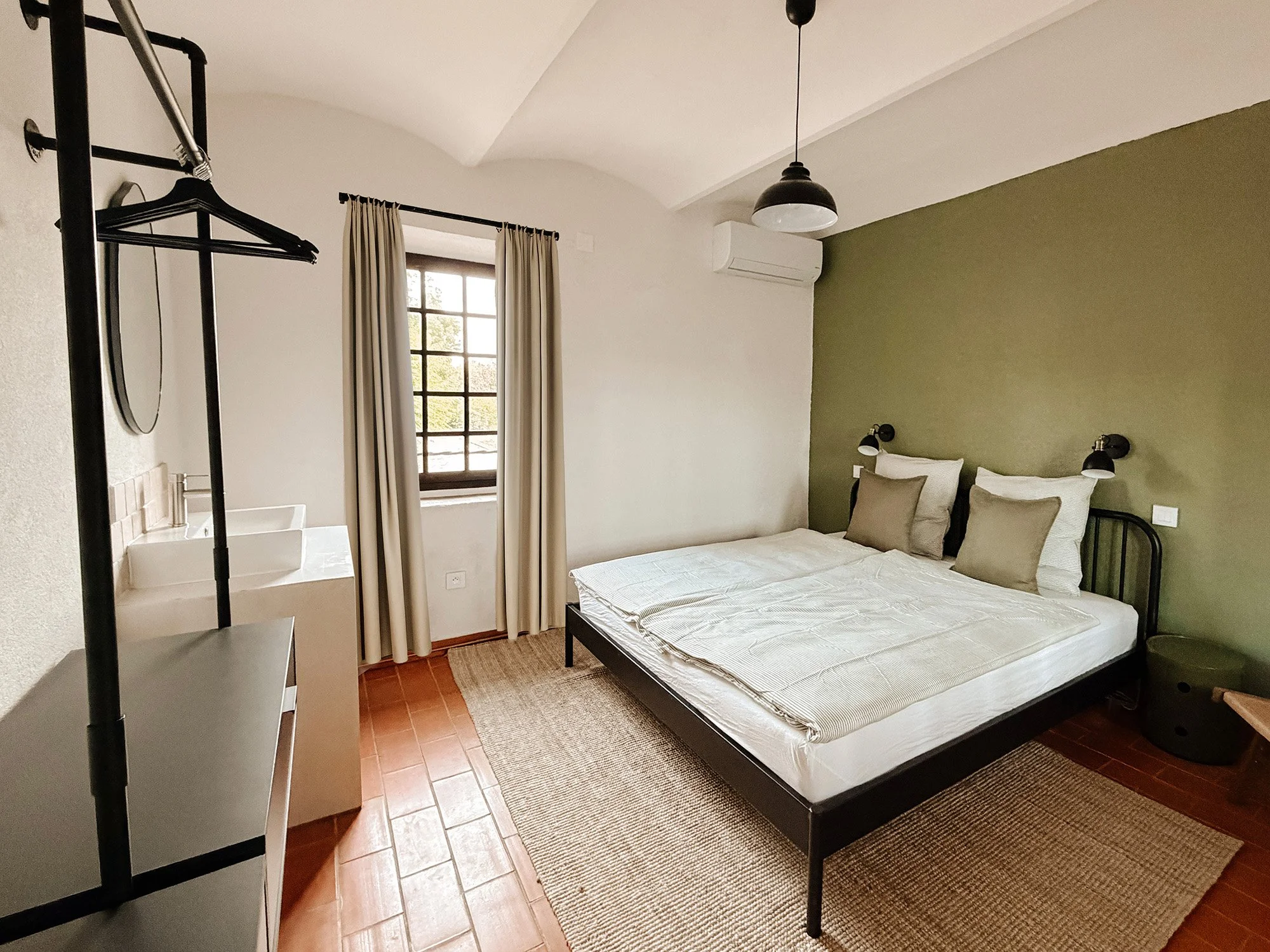 Ein Schlafzimmer mit einem Doppelbett, beige Kissen, einer Stehlampe, einem Fenster mit beige Vorhängen, einer Klimaanlage an der Wand, einem schwarzen Nachttisch und einer kleinen Teppich. Es gibt auch einen Kleiderständer mit Hängern und einen Wasc