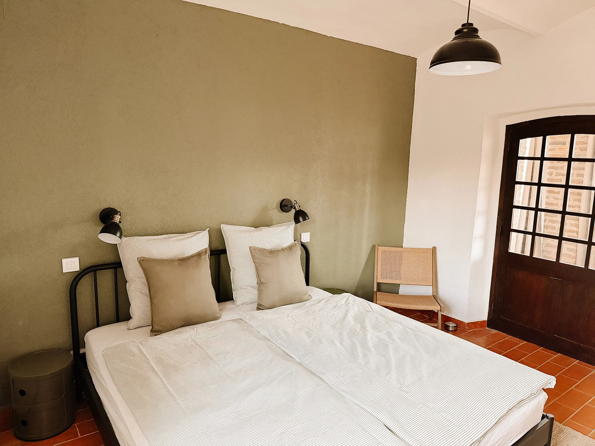 Ein Schlafzimmer mit einem Doppelbett, dezenten Kissen, Nachttischen und Wandlampen, einer, sichtbar gegenüber dem Bett, Ecke mit Stühlen und einem Fenster mit Gitter. Die Wand ist halb grün, halb weiß.