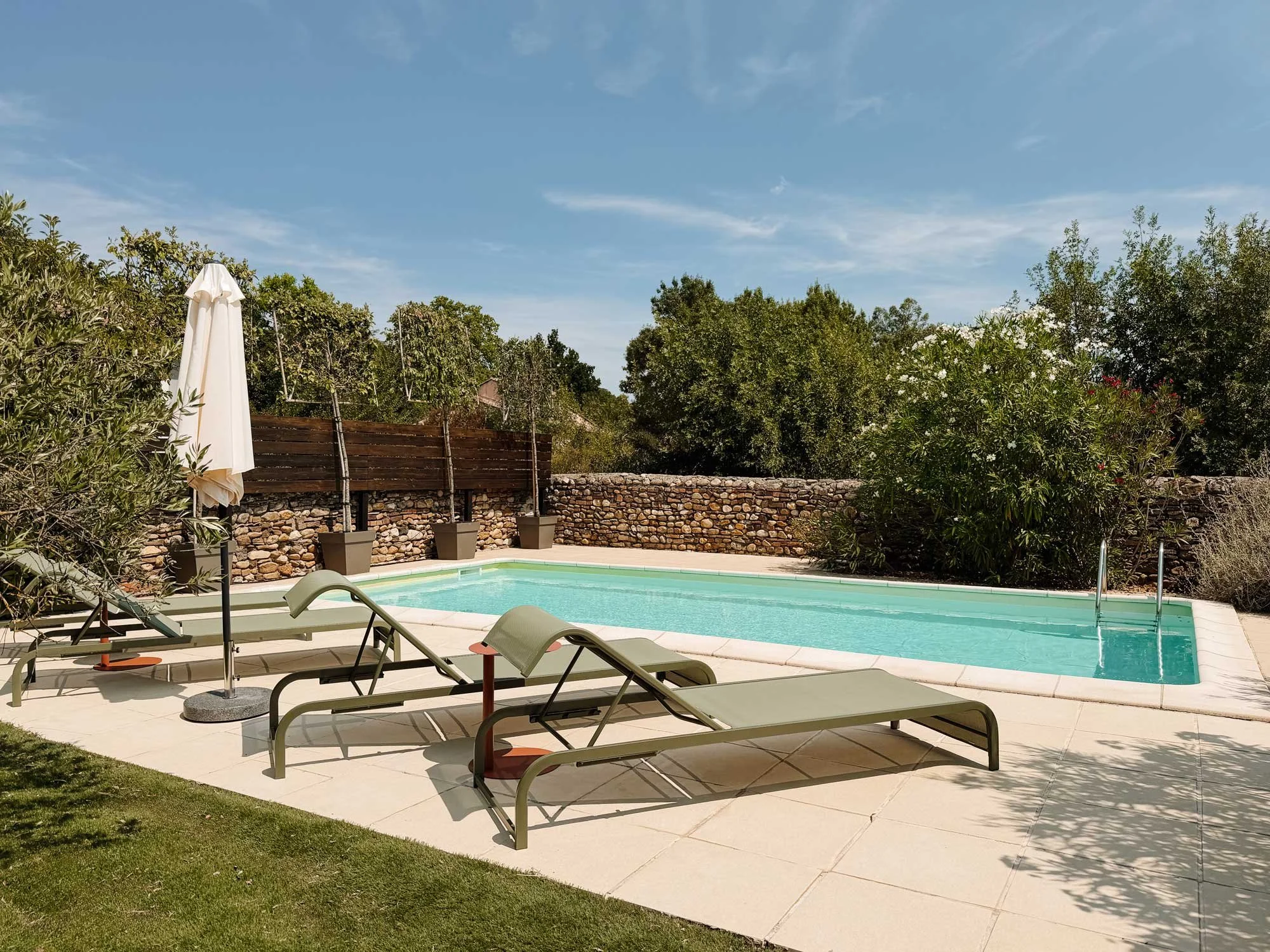 Ein Swimmingpool im Garten mit Liegestühlen und Sonnenschirm, umgeben von Büschen und einer Steinmauer, bei sonnigem Himmel.