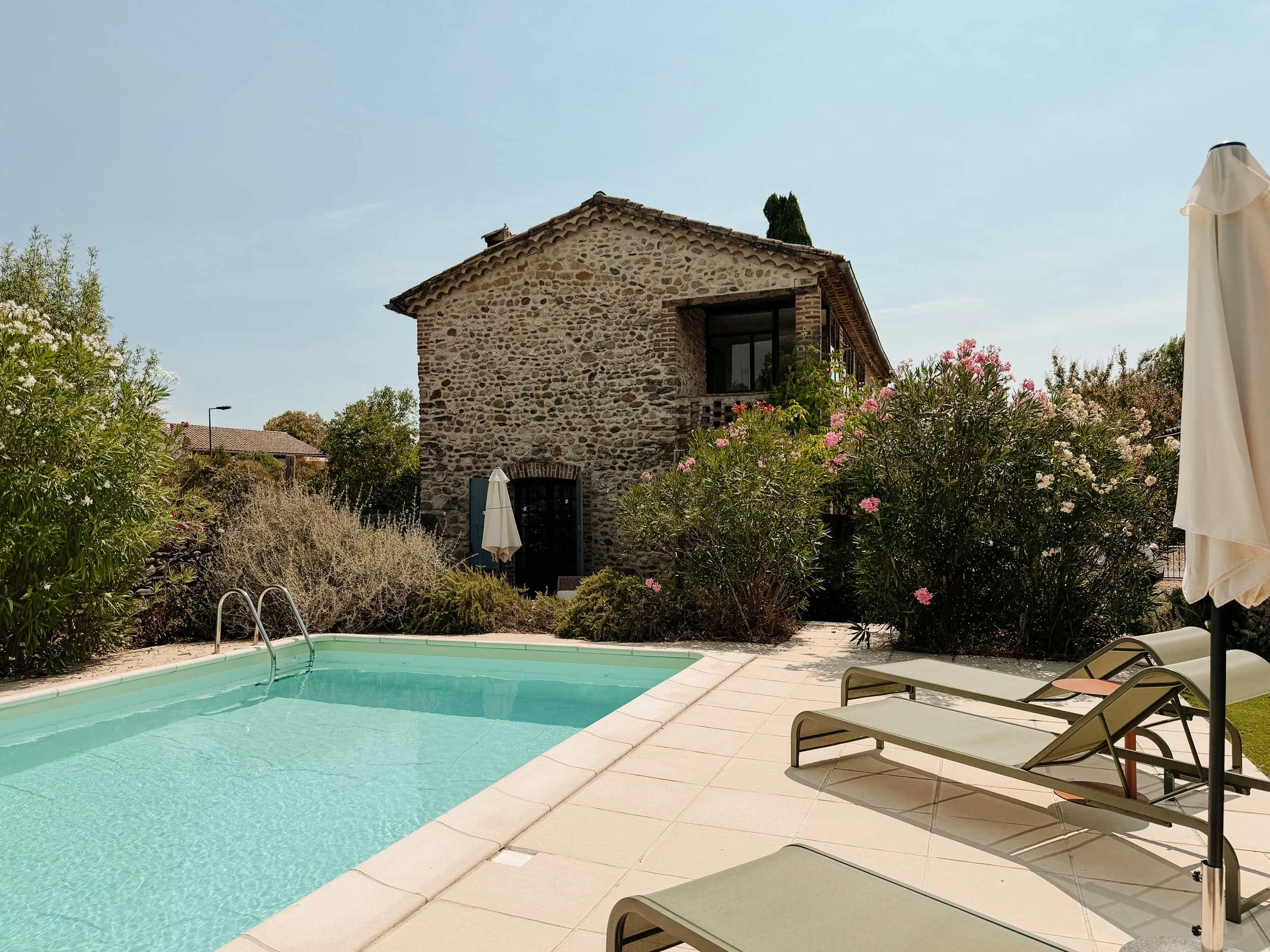 Ein Swimmingpool im Garten neben einem alten Steinhaus mit Fenster und einem Baum im Hintergrund. Es gibt Liegestühle am Pool und eine weiße Sonnenbank.