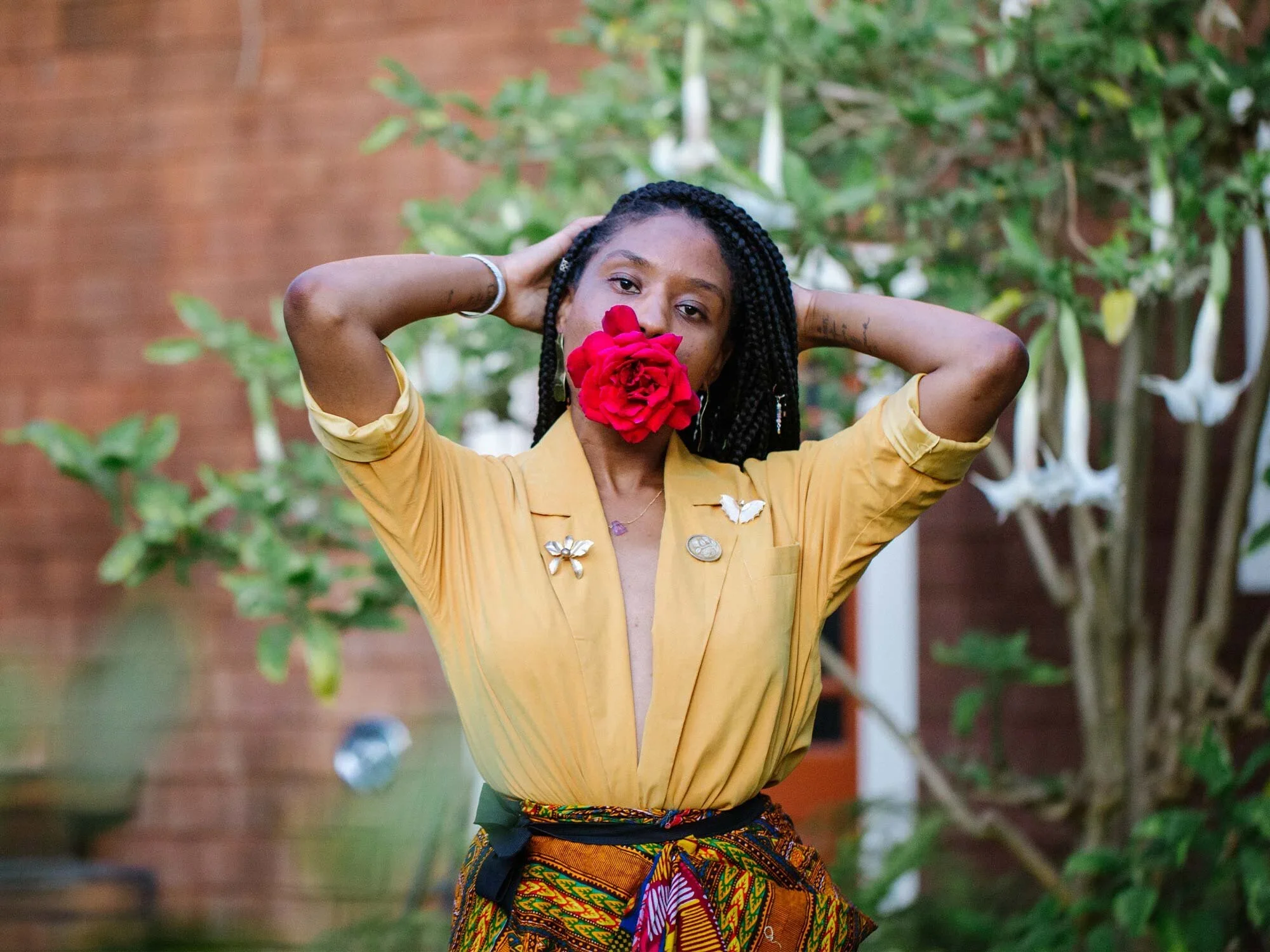 A woman with braided hair holds a red flower in front of her face, with her arms raised and hands behind her head. She is wearing a yellow blouse with decorative pins and a colorful, patterned skirt, standing outdoors with green plants and a brick wall in the background.