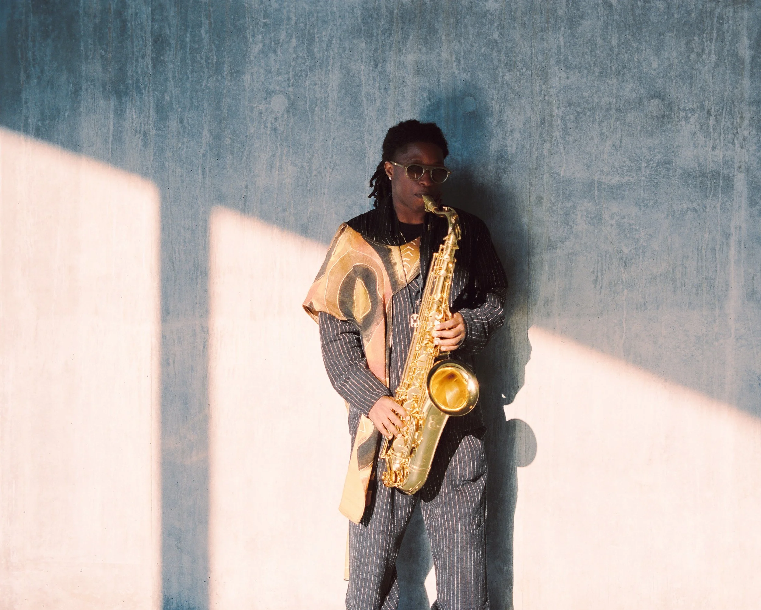 A person with dark brown skin, wearing sunglasses and dressed in a pinstripe suit, playing a saxophone against a textured blue wall with shadows.