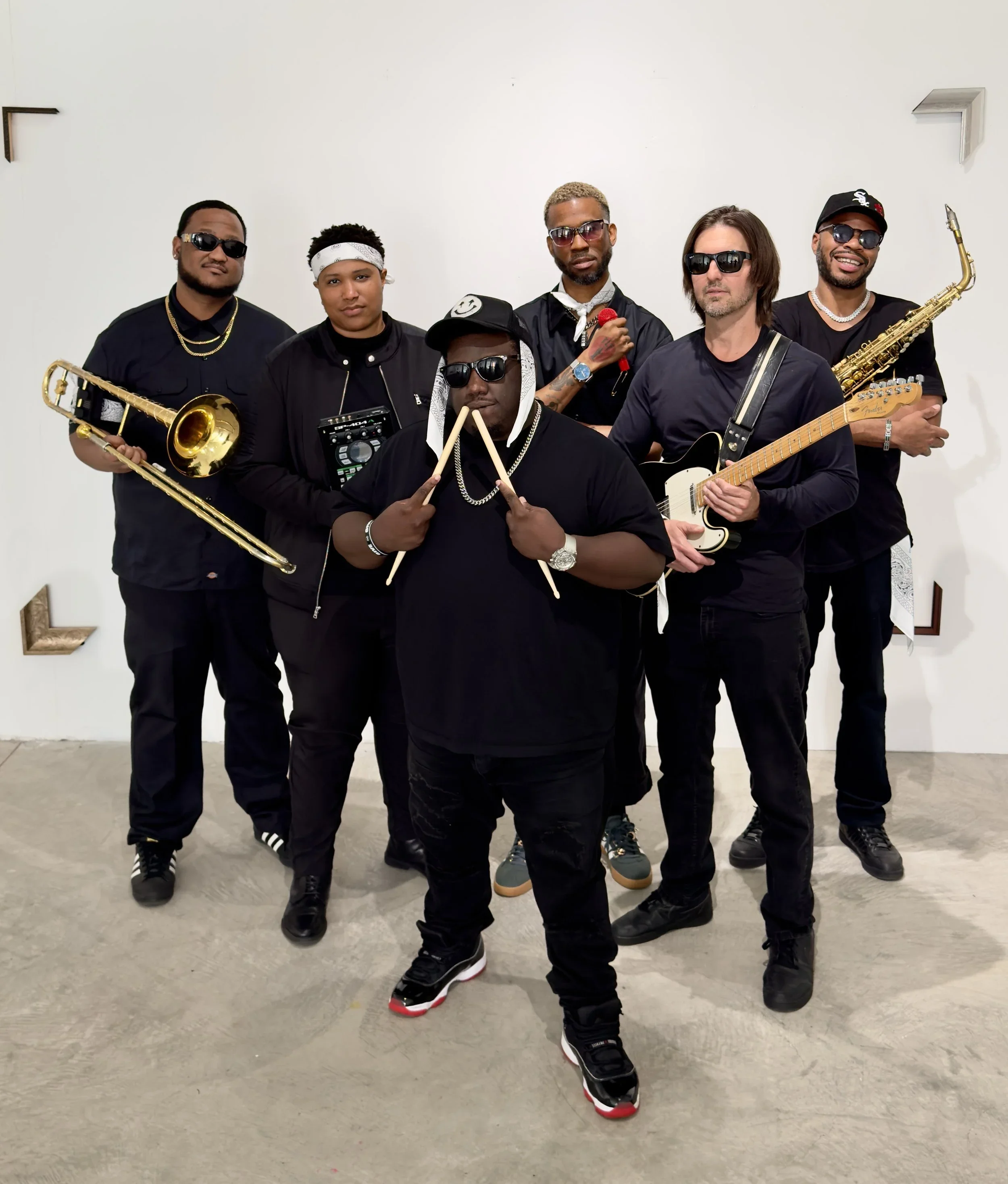 A group of six men posing for a photo against a plain white wall, dressed in casual streetwear, with some holding musical instruments like a saxophone, guitar, and a drum pad, and wearing sunglasses.