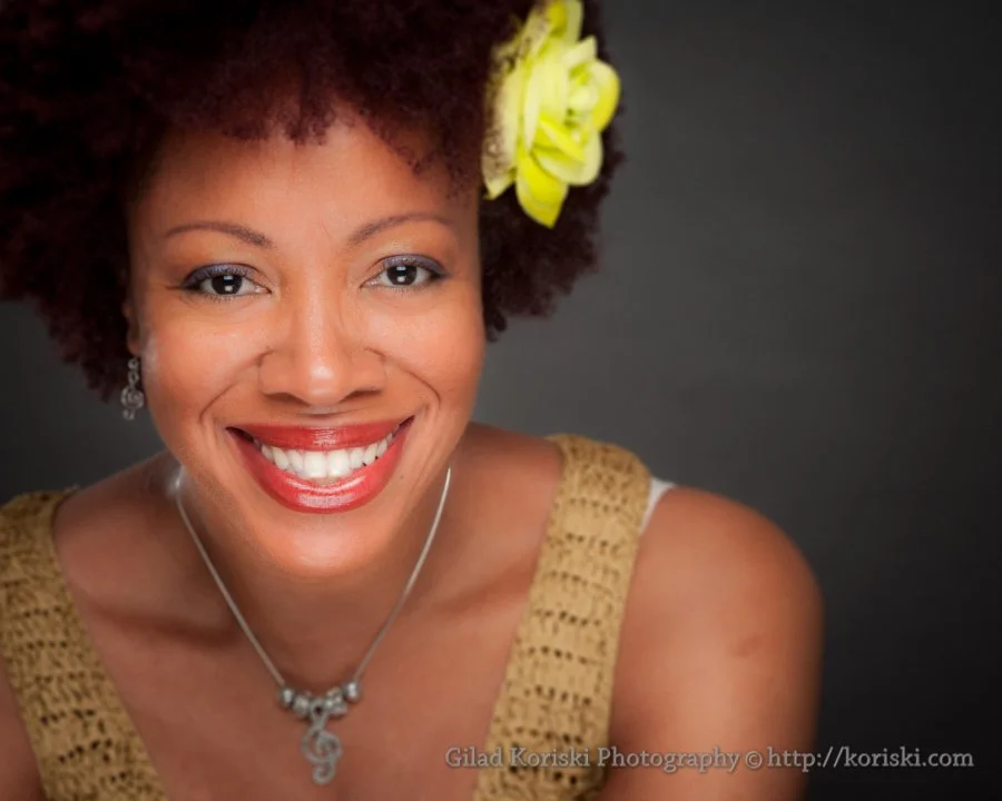 A woman with a big smile, wearing a yellow crocheted top, silver earrings, a treble clef necklace, and a yellow flower in her curly hair against a dark background.