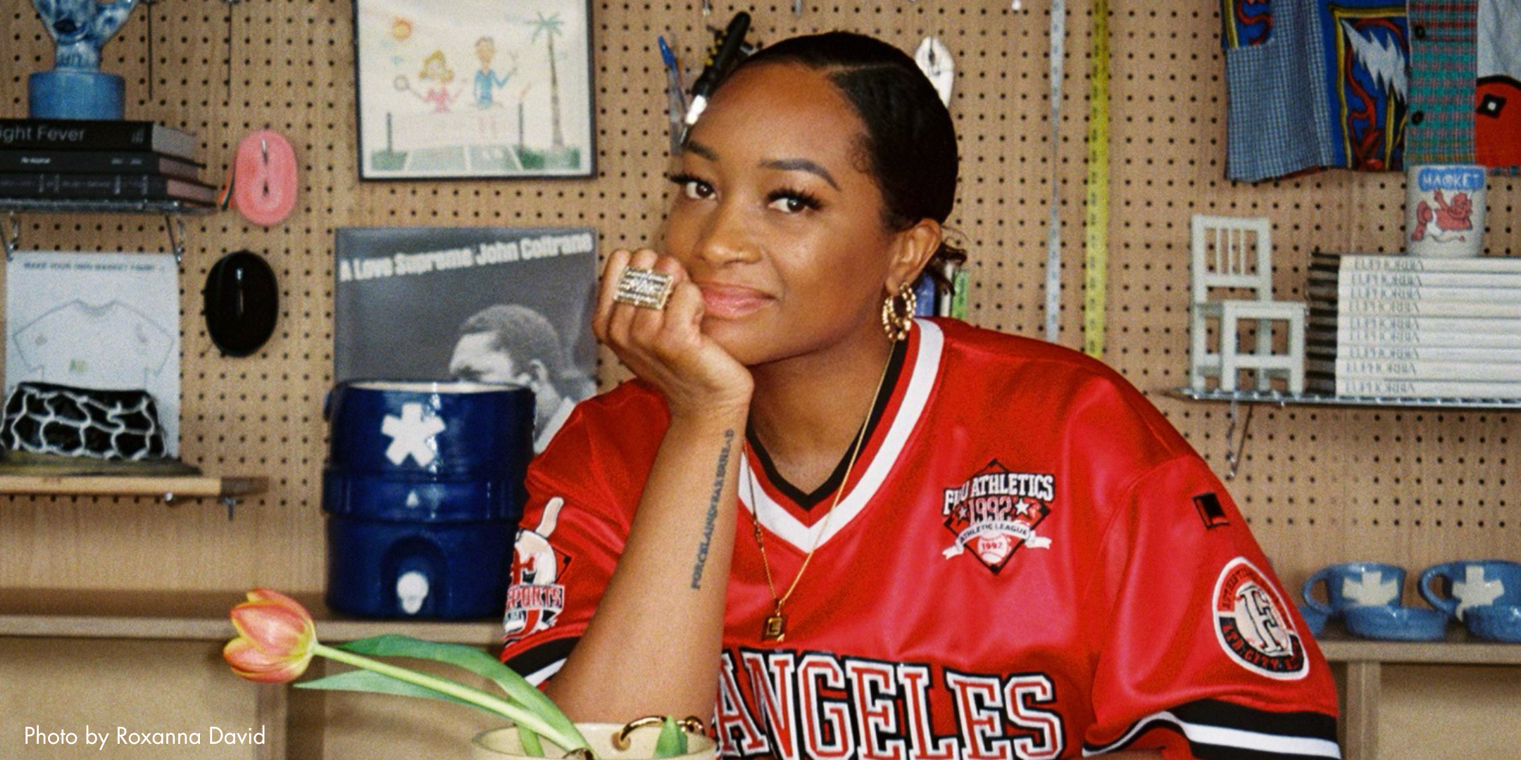 A woman with short hairstyle sitting at a desk, wearing a red sports jersey with 'Los Angeles' written on it, in a room with miscellaneous items on shelves and a pegboard background.