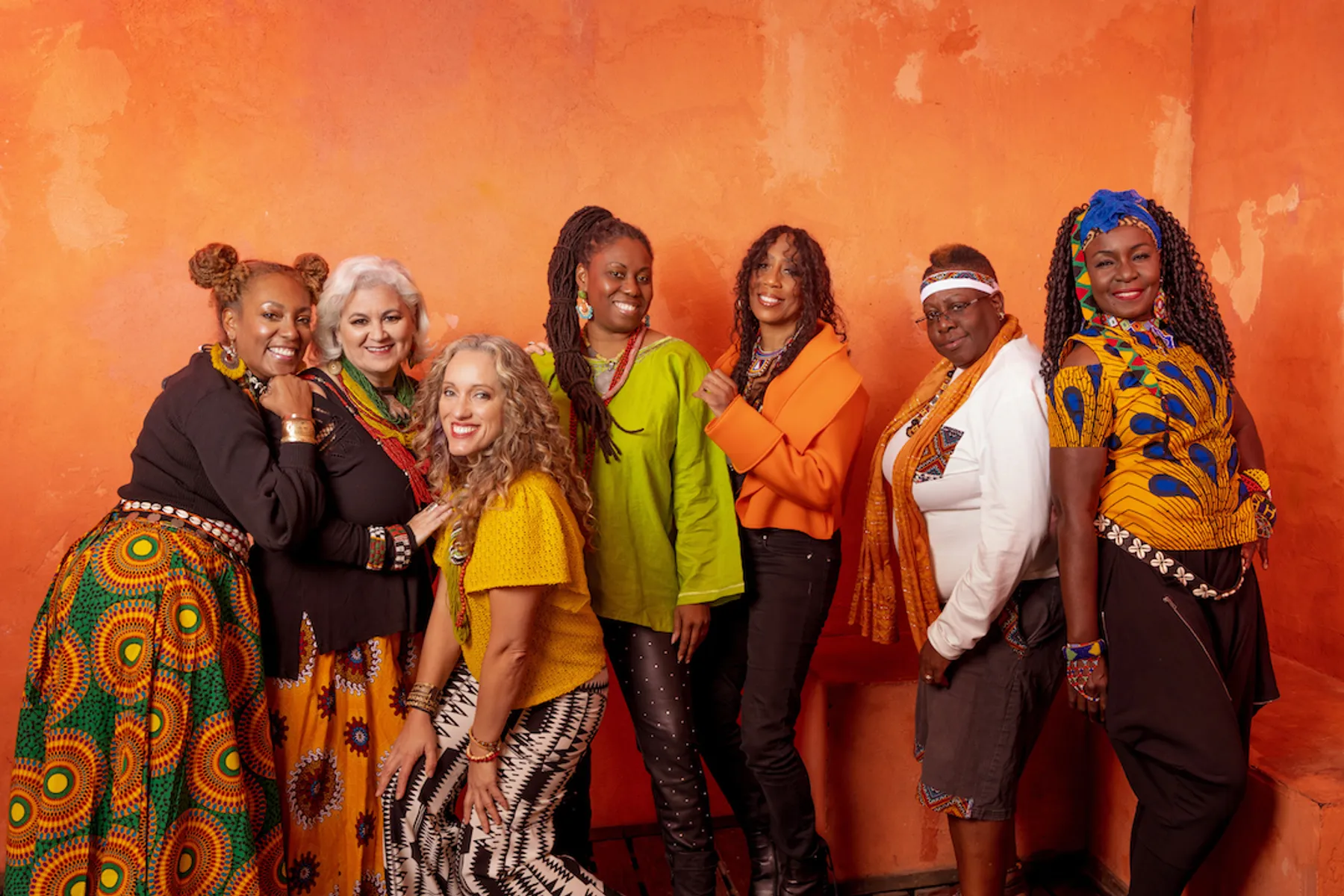 A diverse group of eight women standing together against a textured orange wall, wearing colorful traditional and modern clothing, smiling at the camera.