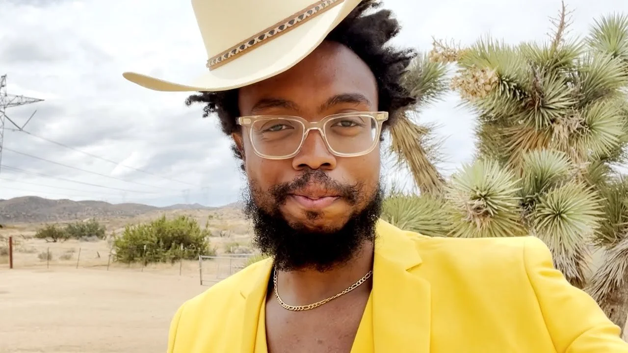 A man with dark curly hair, beard, and glasses taking a selfie outdoors. He is wearing a yellow jacket, a gold chain, and a beige cowboy hat. The background shows desert landscape with a yucca plant, some bushes, hills, and power lines under a cloudy sky.