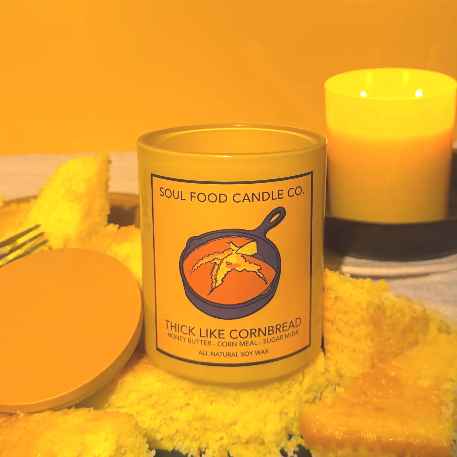 A yellow candle from Soul Food Candle Co. labeled 'Thick Like Cornbread' with honey butter, corn meal, and sugar musk scent, alongside another lit candle on a black plate, surrounded by yellow cornmeal.
