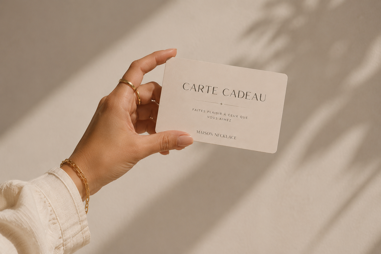 Main dans une main tenant une carte cadeau blanche avec des écritures en français, portant des bijoux en or, avec un fond lumineux et ombré.
