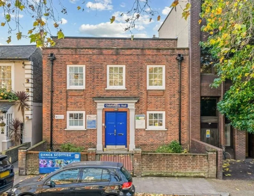 A brick building with a blue double door entrance labeled "Oddfellows Hall," four windows, and surrounded by trees and parked cars.